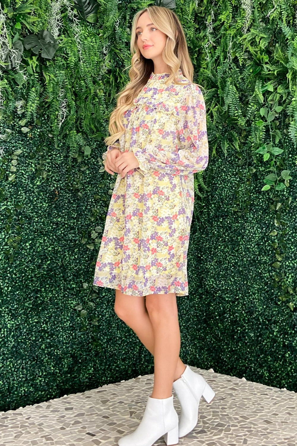 A woman stands in front of a leafy green background wearing the And The Why Floral Mock Neck Flounce Sleeve Dress, smiling and looking to the side.