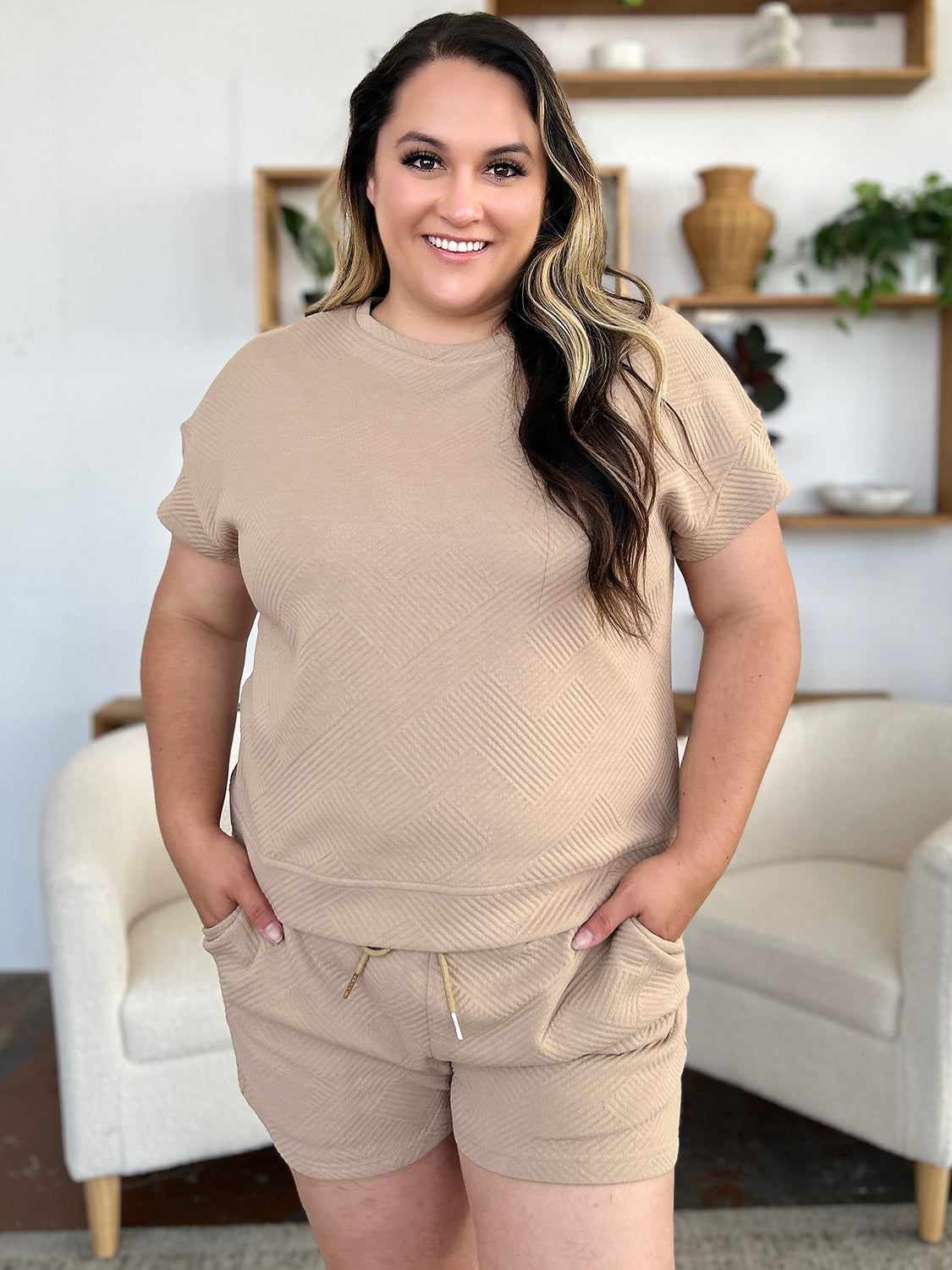 A woman with long dark hair is wearing the Double Take Full Size Texture Short Sleeve T-Shirt and Drawstring Shorts Set, made from beige polyester. The set features a short-sleeve top and matching shorts, with the woman standing confidently with one hand in her pocket and maintaining a neutral expression. For easy care, remember to machine wash cold.