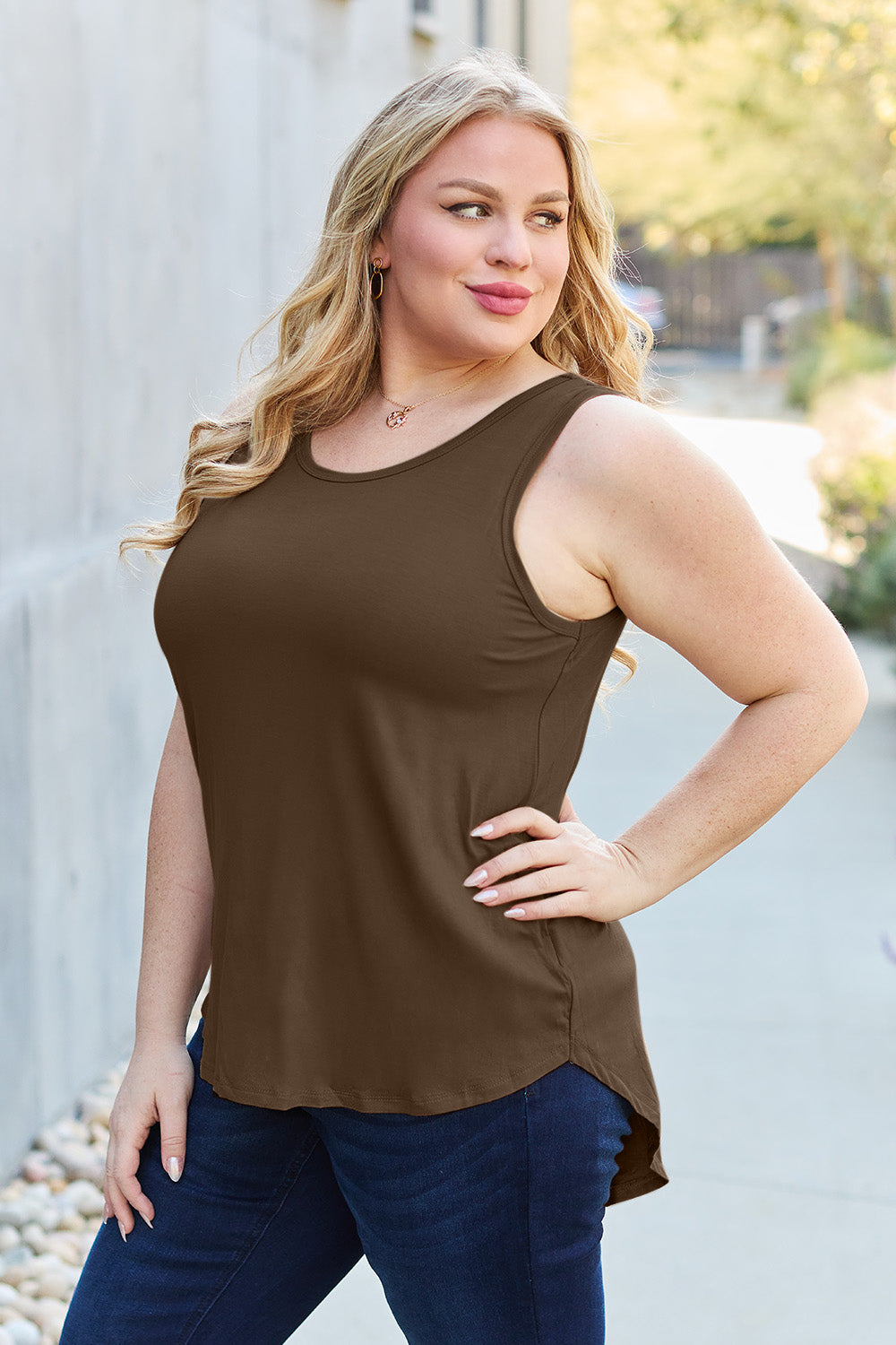 A woman wearing sunglasses, blue jeans, and the Basic Bae Full Size Round Neck Curved Hem Tank is standing in front of a concrete wall, holding her glasses with one hand and carrying a beige shoulder bag. Her basic style complements the slightly stretchy fabric of her outfit, ensuring comfort while maintaining chic simplicity.