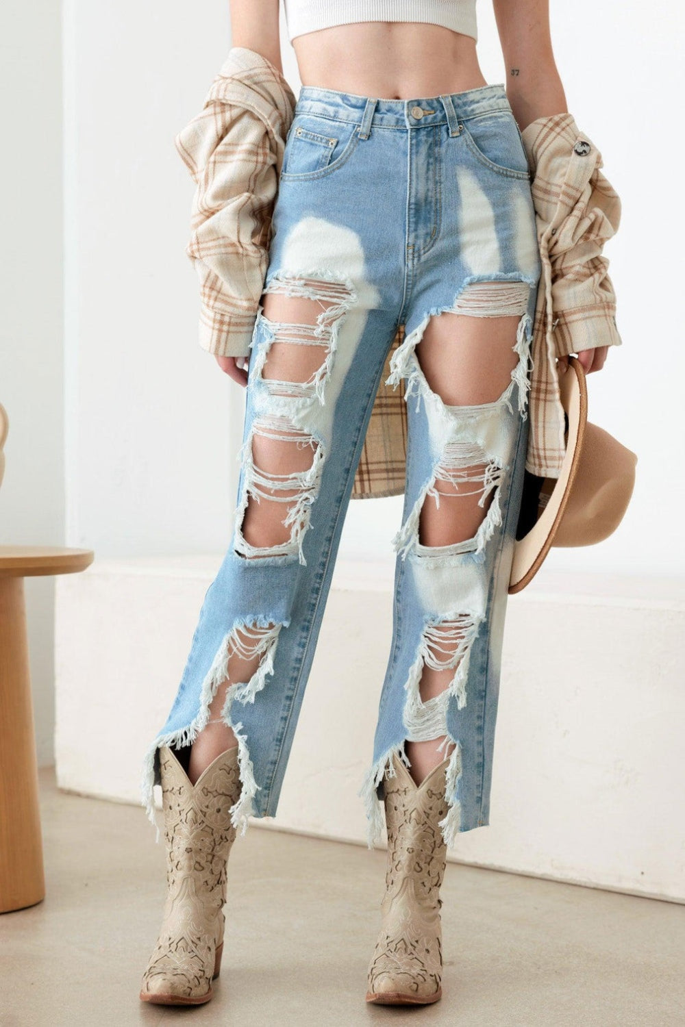 A woman stands indoors in a Western Boho style, showcasing a white crop top and plaid shirt paired with the Litz La Frayed Cut Distressed Jeans. She holds a hat in her hand and completes her outfit with patterned boots, while a vase is positioned on a wooden stool nearby.