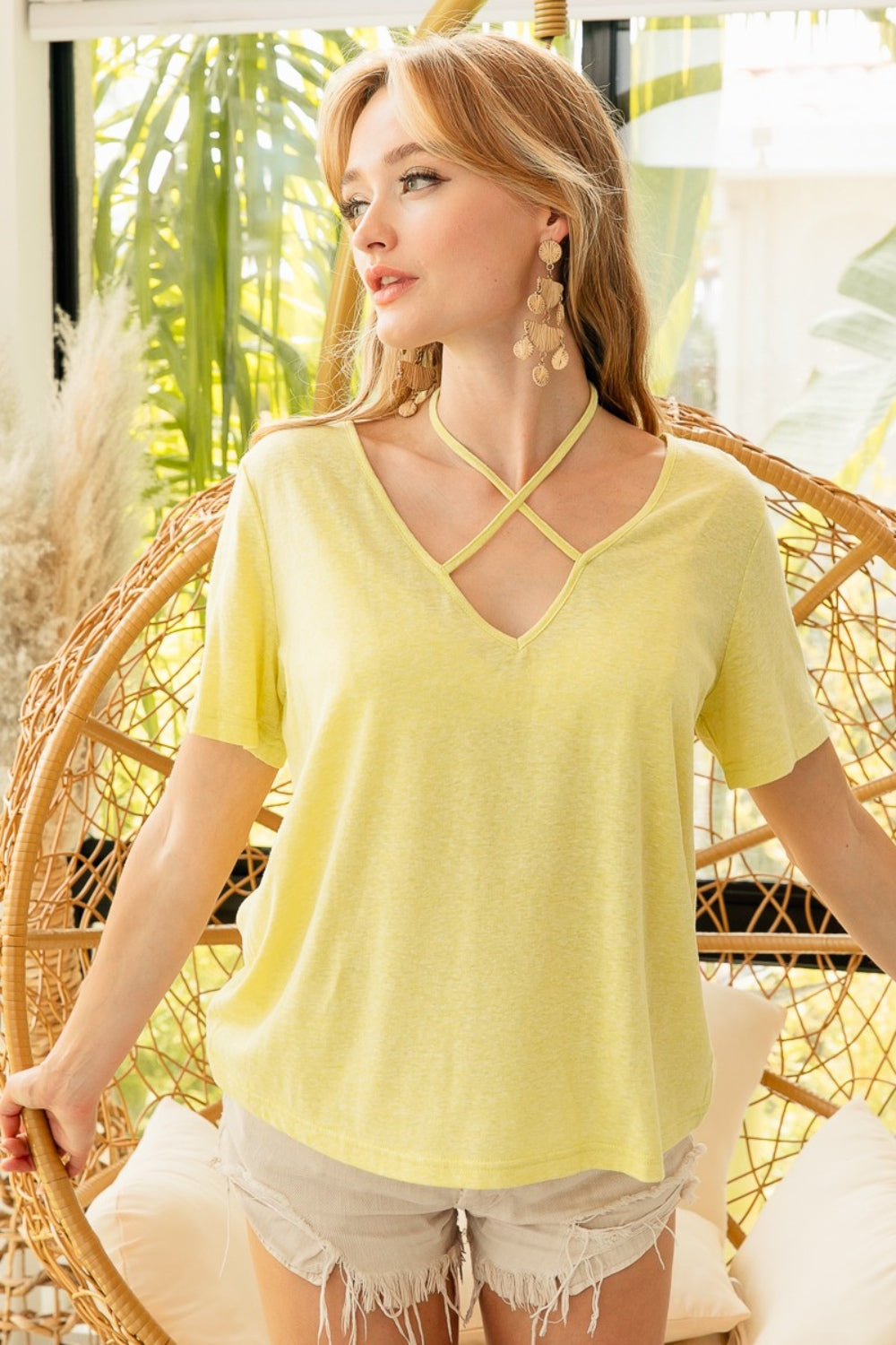 A person in a trendy yellow BiBi Tied Detail Short Sleeve T-Shirt and beige shorts stands in front of a wicker chair in a bright room.