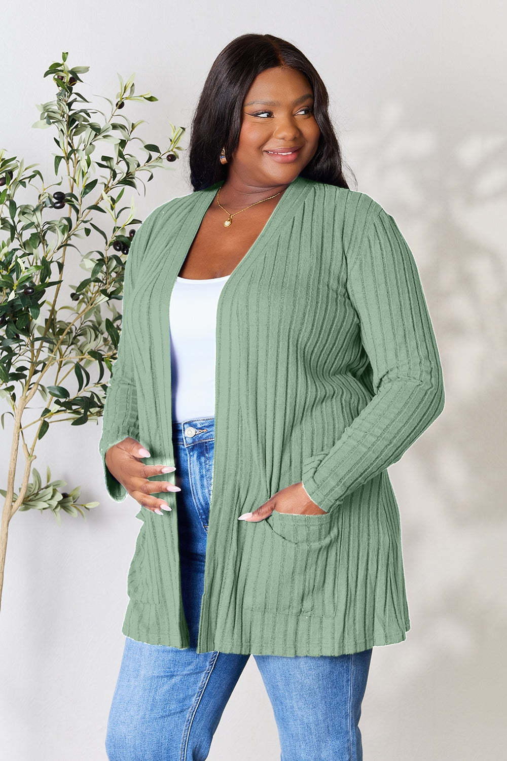 A woman in a Basic Bae Full Size Ribbed Open Front Cardigan with Pockets and jeans, made from stretchy material, stands in front of a plant, smiling slightly and looking to the side.