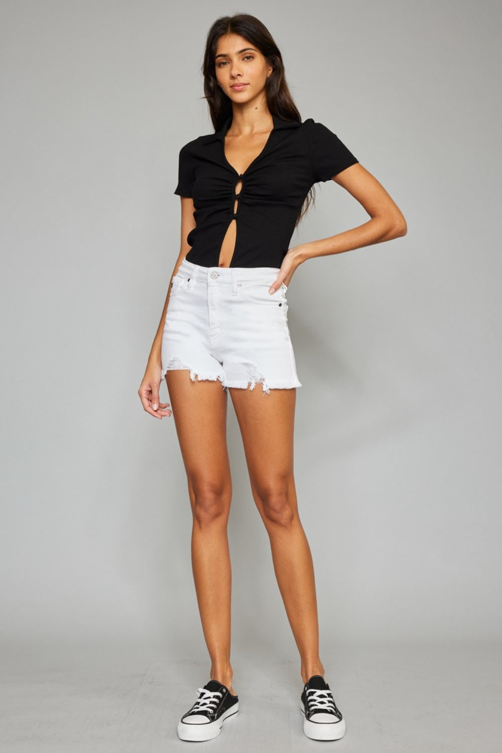 A woman with long dark hair is wearing a black short-sleeve top with cut-out details and the Kancan Full Size High Rise Frayed Hem Denim Shorts. She is standing against a plain gray background, showcasing her chic summer wardrobe.