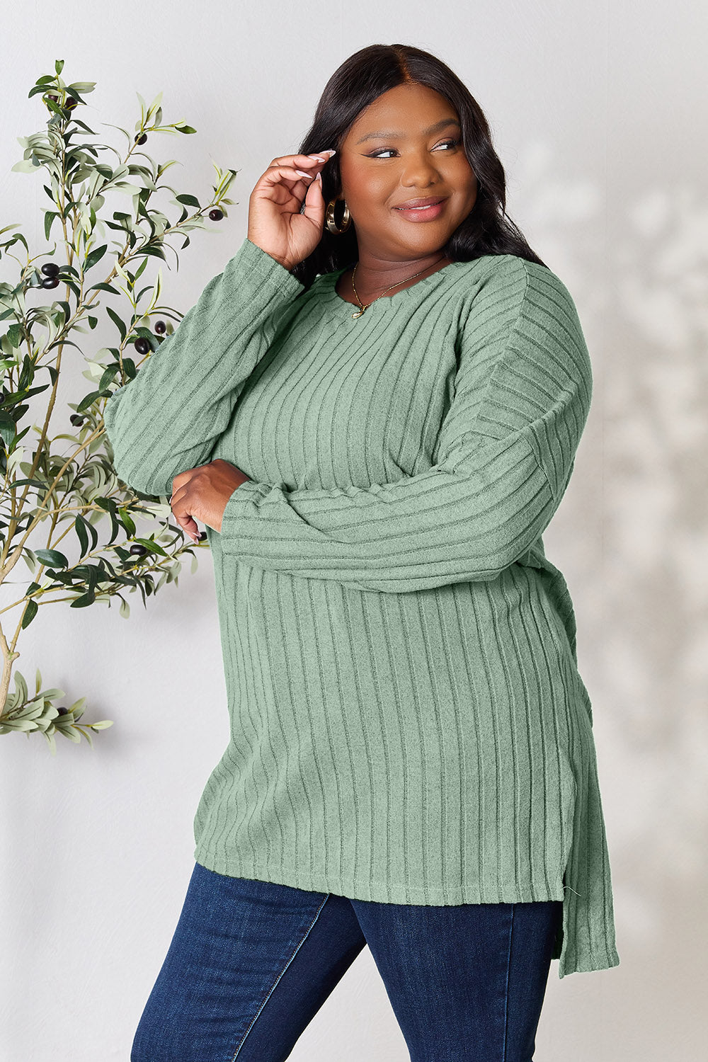 A person with shoulder-length hair smiles while wearing a beige ribbed sweater called the Basic Bae Full Size Ribbed Round Neck Long Sleeve Slit Top, along with matching beige pants, showcasing a basic style. They stand next to a green plant.