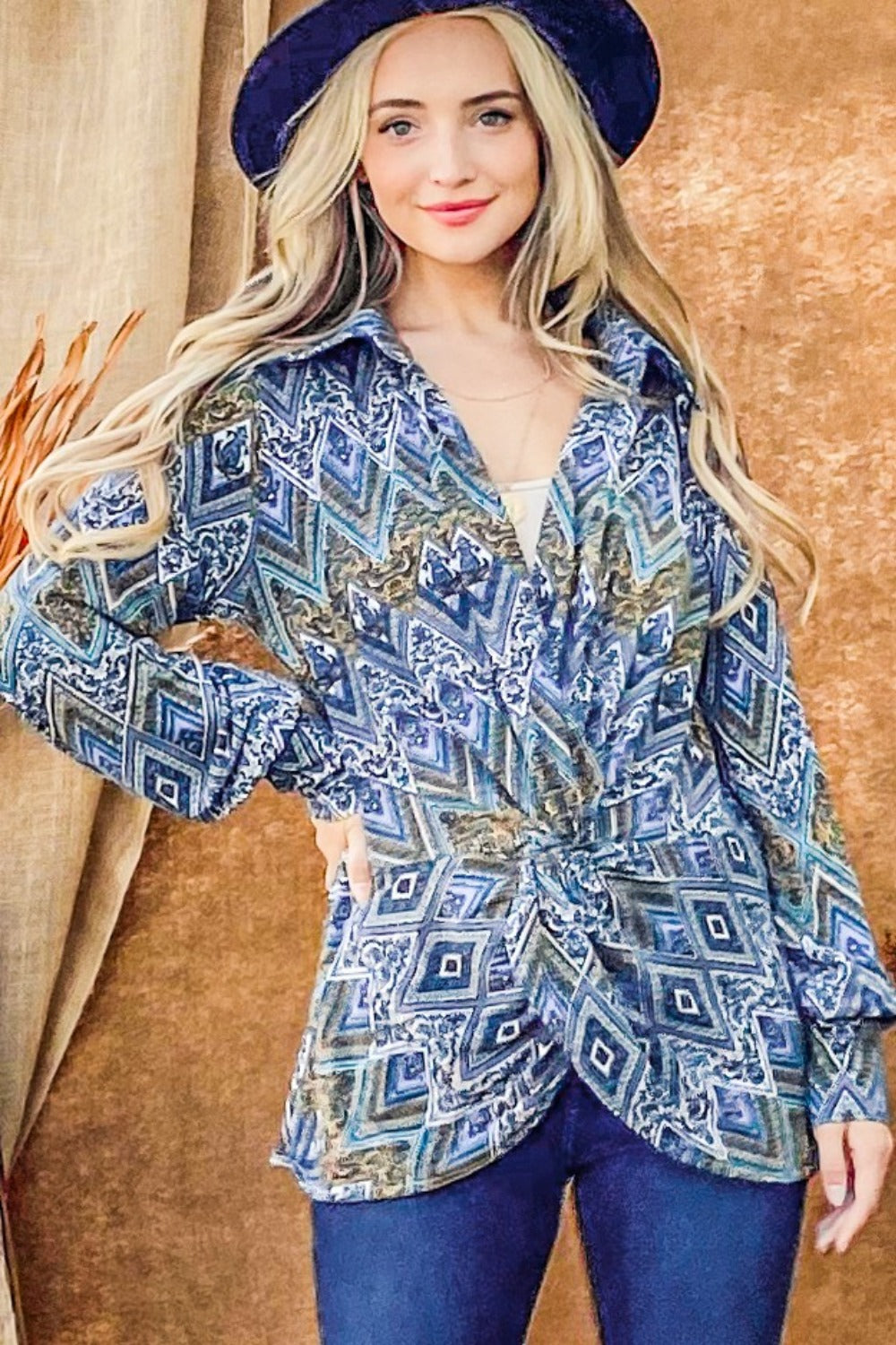 A woman with long blonde hair wearing an And The Why Print Twist Knot Long Sleeve Blouse, blue jeans, and a dark blue hat stands in front of a brown backdrop.