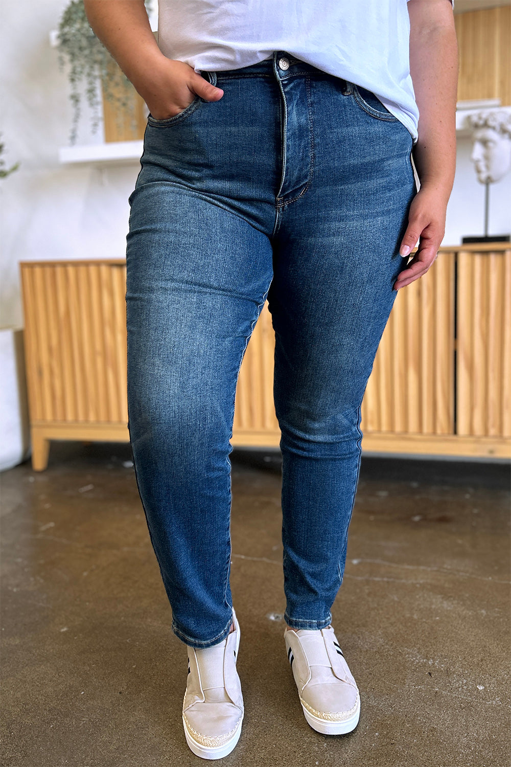 Person wearing Judy Blue Full Size Tummy Control High Waist Slim Jeans in blue and a white shirt, with their hands in their pockets. They are standing indoors on a polished concrete floor with a wooden cabinet and plant in the background.