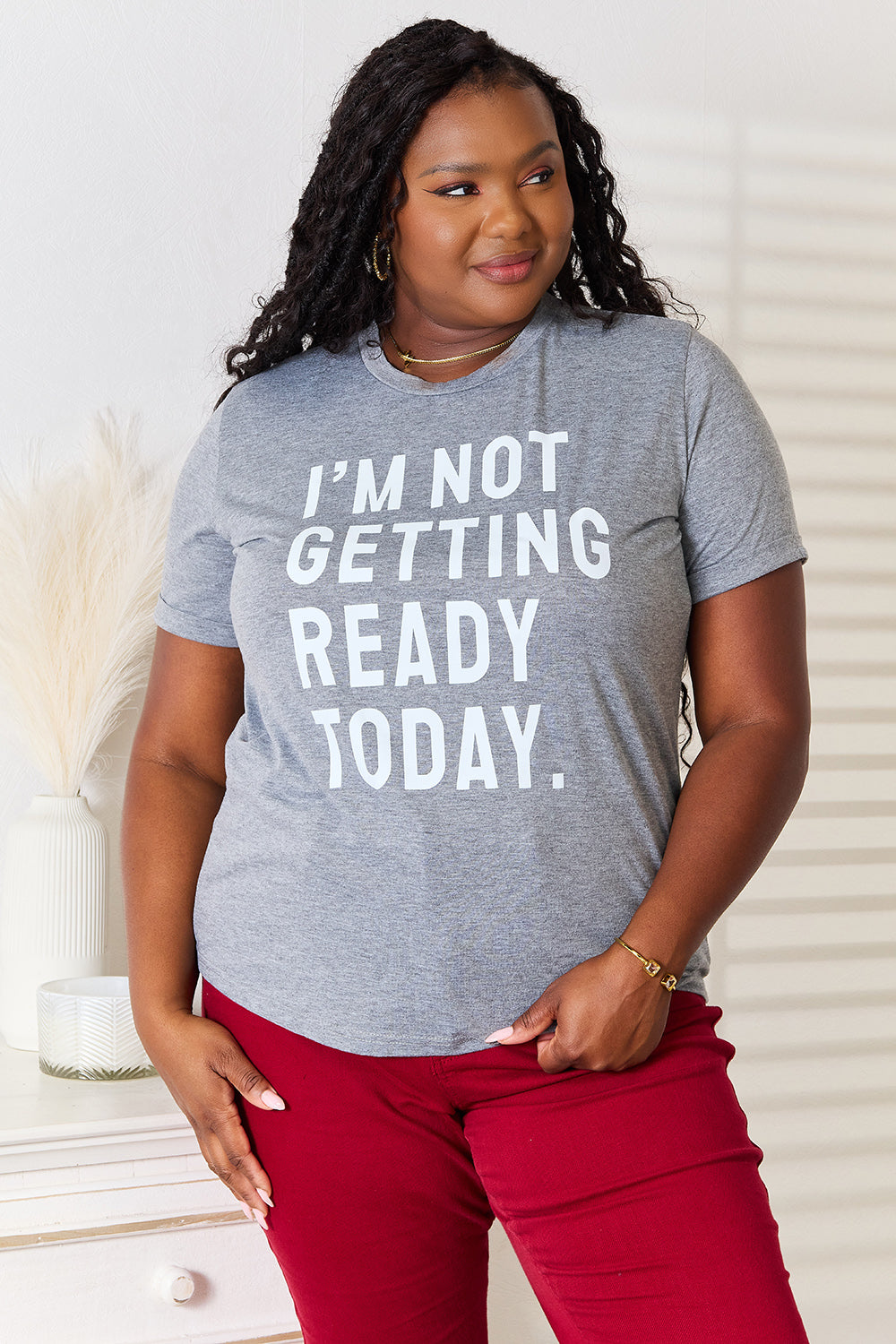 Dressed in a Simply Love I'M NOT GETTING READY TODAY Graphic T-Shirt and vibrant red pants, the individual exudes a carefree style while standing near a white surface adorned with decorative objects.