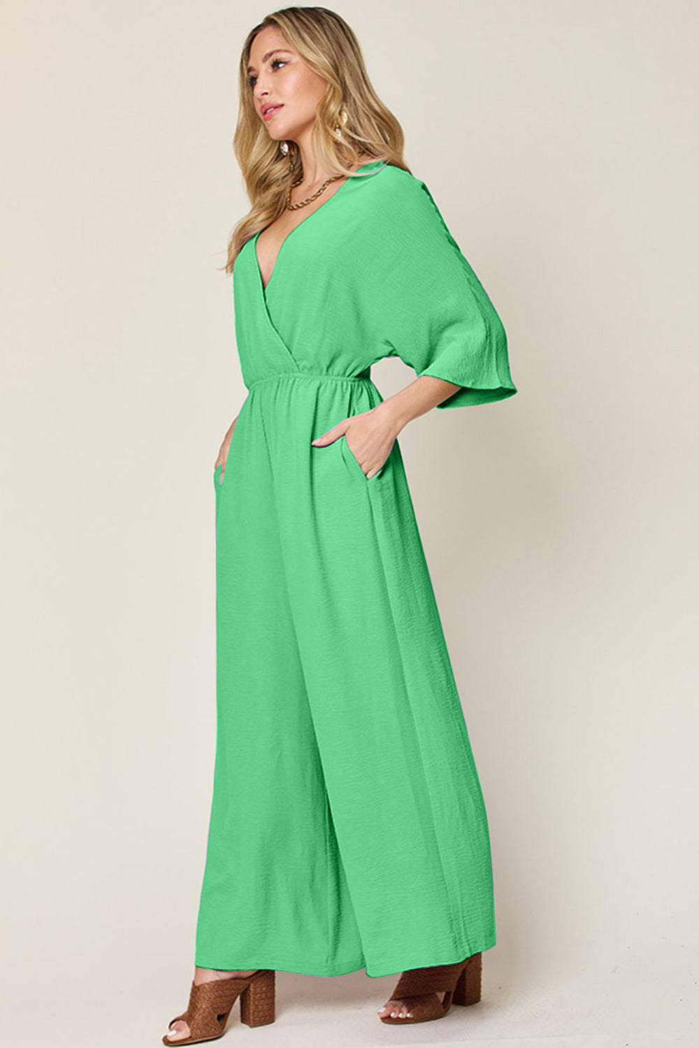 Person wearing the Double Take Full Size Surplice Wide Leg Jumpsuit with Pockets in royal blue, featuring wide legs, a deep V-neckline, and ruched detailing, posing with hands on hips against a plain light-colored background.