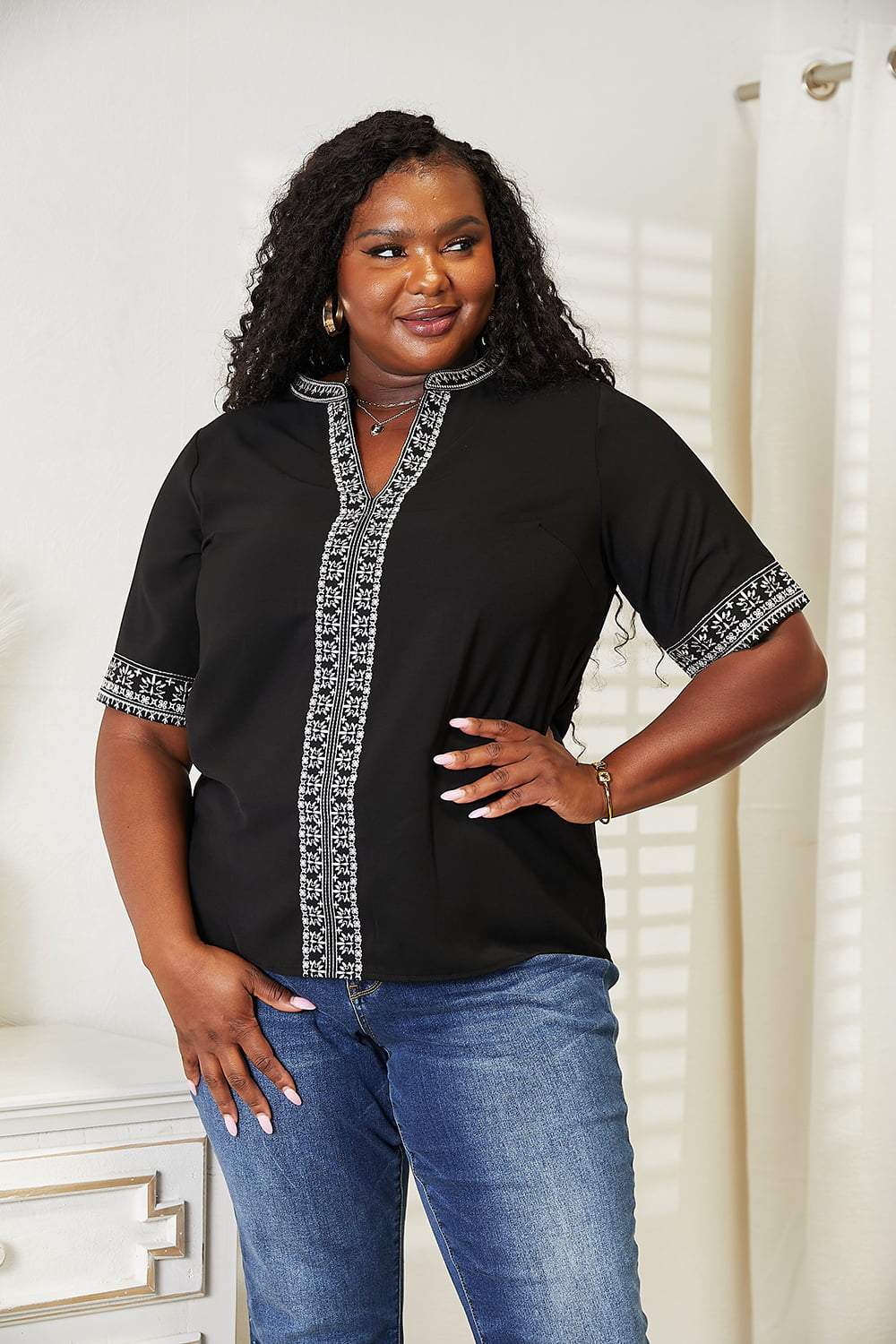 Wearing the elegant Double Take Embroidered Notched Neck Top and blue jeans, a woman with long, wavy hair stands smiling against a light background.