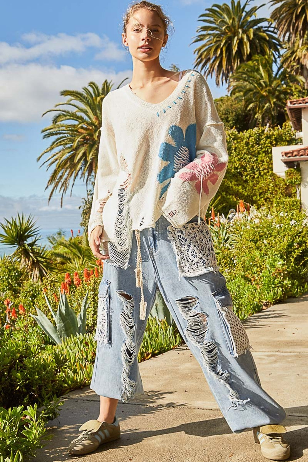 A person with curly blonde hair is standing outside, smiling. They are wearing a stylish and edgy POL Distressed Flower V-Neck Dropped Shoulder Knit Top. Palm trees and other foliage are in the background.