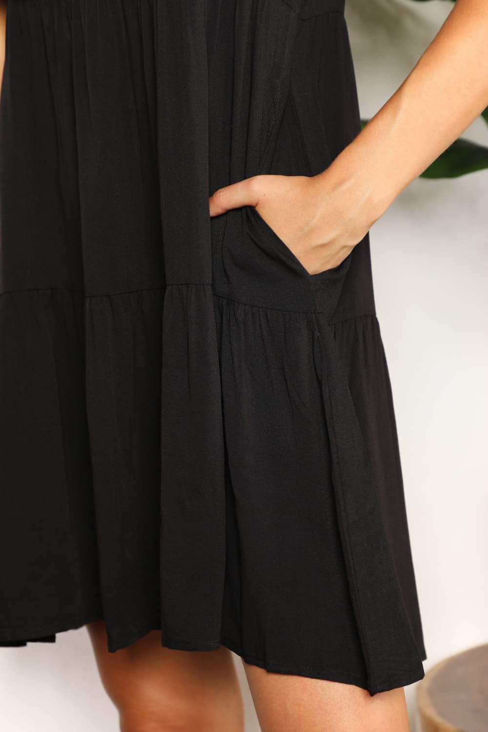 A woman stands indoors wearing the Mandy V-Neck Flounce Sleeve Tiered Dress in navy blue. She has one hand on her hip and is smiling.