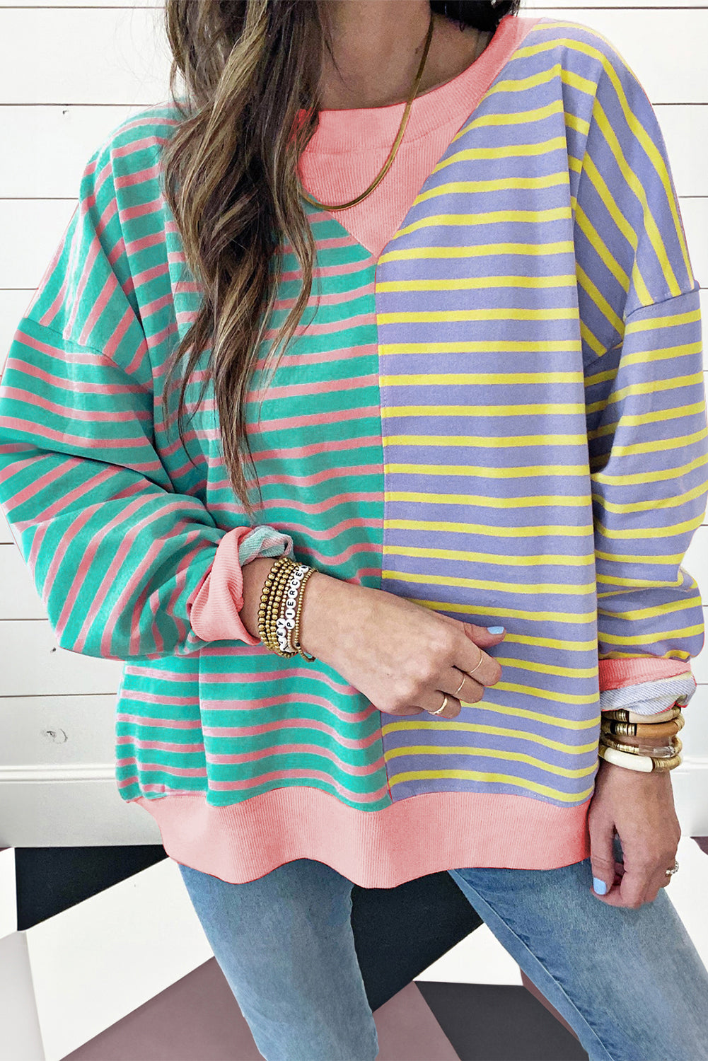 A person with wavy hair is seen from behind wearing a Casual Stripe Colorblock Drop Shoulder Oversize Sweatshirt in pastel colors and blue jeans. They are carrying a white shoulder bag, showcasing a trendy color block design.
