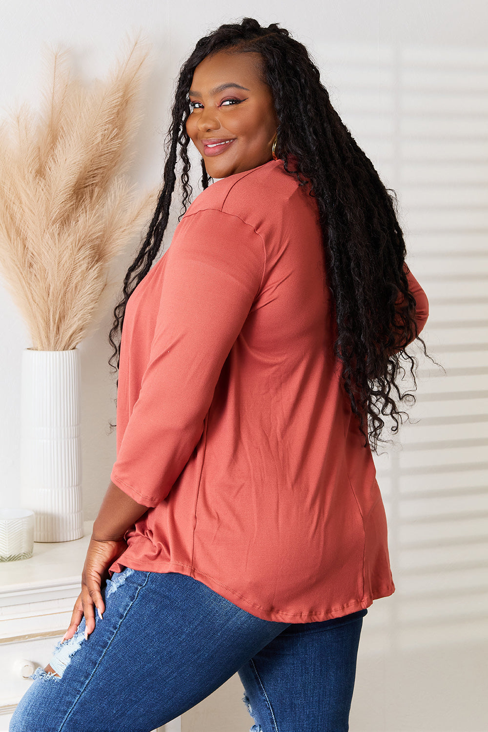 A person with long hair is wearing the Culture Code Full Size Open Front Cardigan in a rust color, complemented by a flattering draped front. They pair it with a white top and ripped jeans while standing indoors with one hand on their hip.