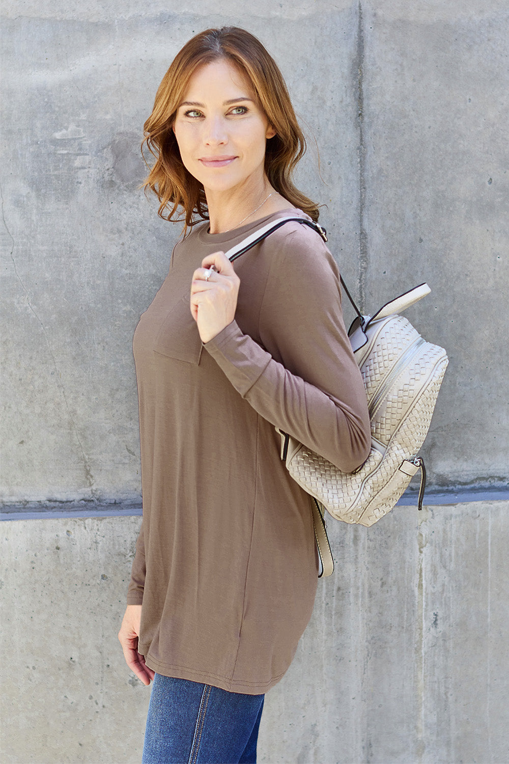 A woman with long hair wearing sunglasses, a Basic Bae Full Size Round Neck Long Sleeve Top in turquoise, and jeans is standing in front of a concrete wall with one hand on her hip.