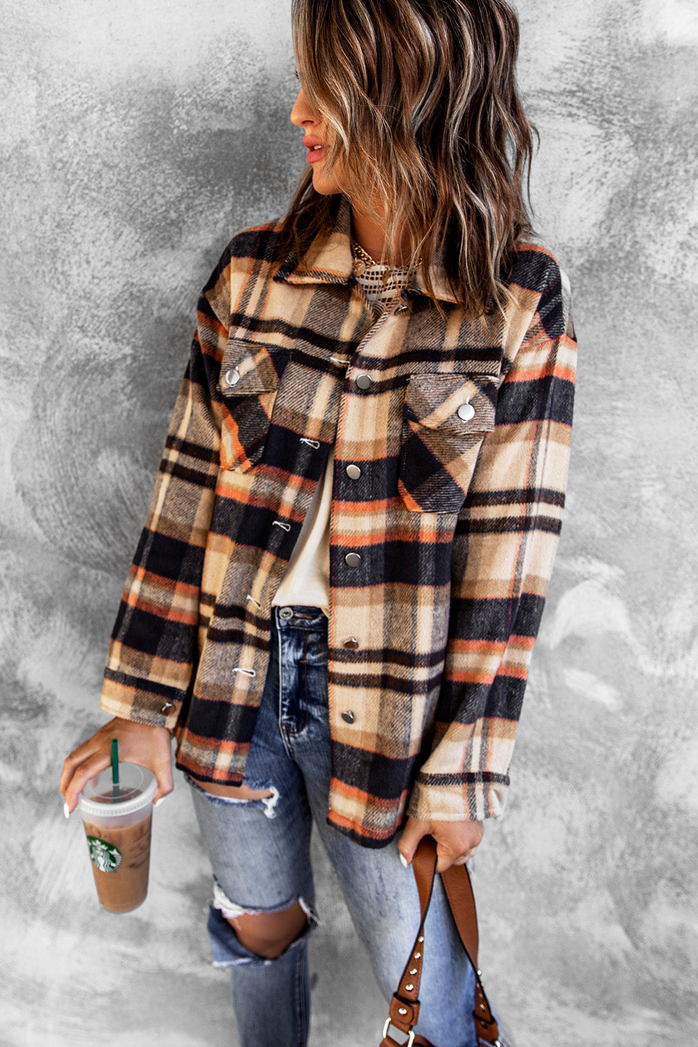 A person wearing a Brown Plaid Button Up Long Sleeve Flannel Shacket layered over a white top with ripped jeans and holding a brown handbag stands against a textured gray wall.