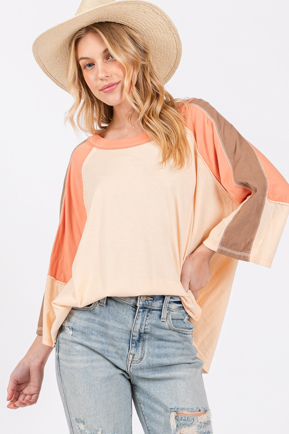 A woman dons a wide-brimmed hat and a modern look with her SAGE + FIG Color Block Curved Hem T-Shirt paired effortlessly with jeans, creating a versatile piece that showcases her vibrant style.