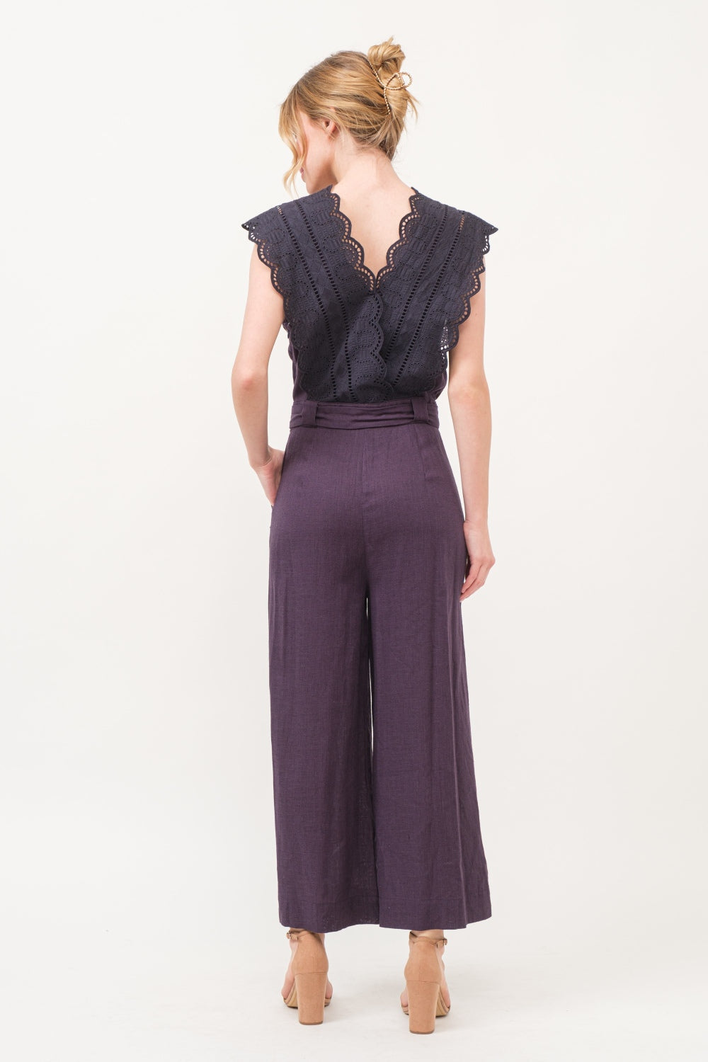 A person in an elegant and fashionable And The Why Laced Surplice Tie Waist Jumpsuit with wide-leg pants stands against a plain background, smiling with hands on hips.