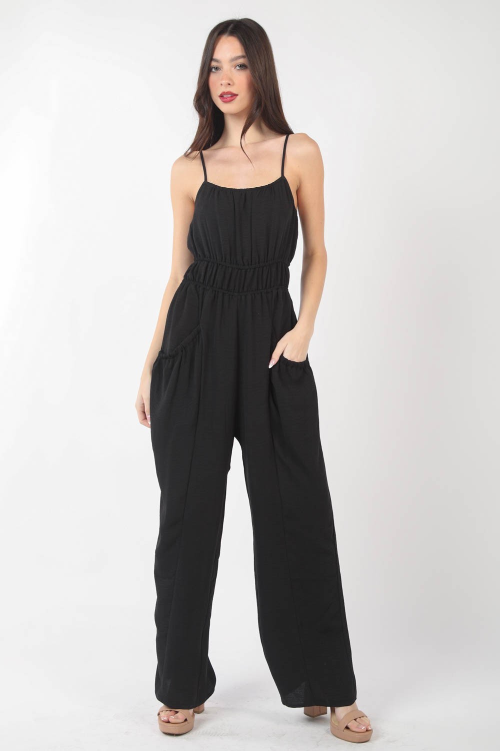 A woman stands against a plain backdrop wearing the VERY J Pintuck Detail Woven Sleeveless Jumpsuit. The black jumpsuit has thin straps, wide-leg pants, and is crafted from woven fabric with subtle pintuck details. She has her hands in her pockets and is looking at the camera.