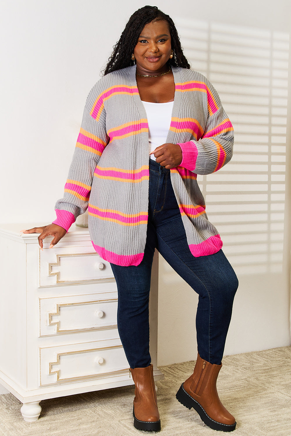 A woman stands against a white wall, smiling with one hand on her hip. She wears the Woven Right Ribbed Long Sleeve Cardigan, featuring pink and orange stripes, over a white top and dark jeans—an excellent choice for versatile layering.