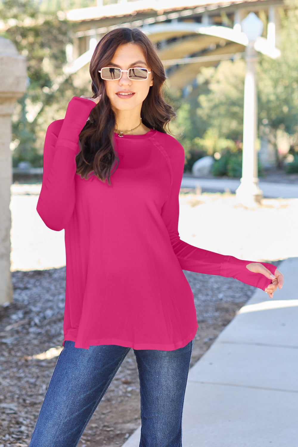 A woman wearing sunglasses and a slightly stretchy Basic Bae Full Size Round Neck Long Sleeve T-Shirt in white stands on a sidewalk with an arched structure in the background, smiling slightly.