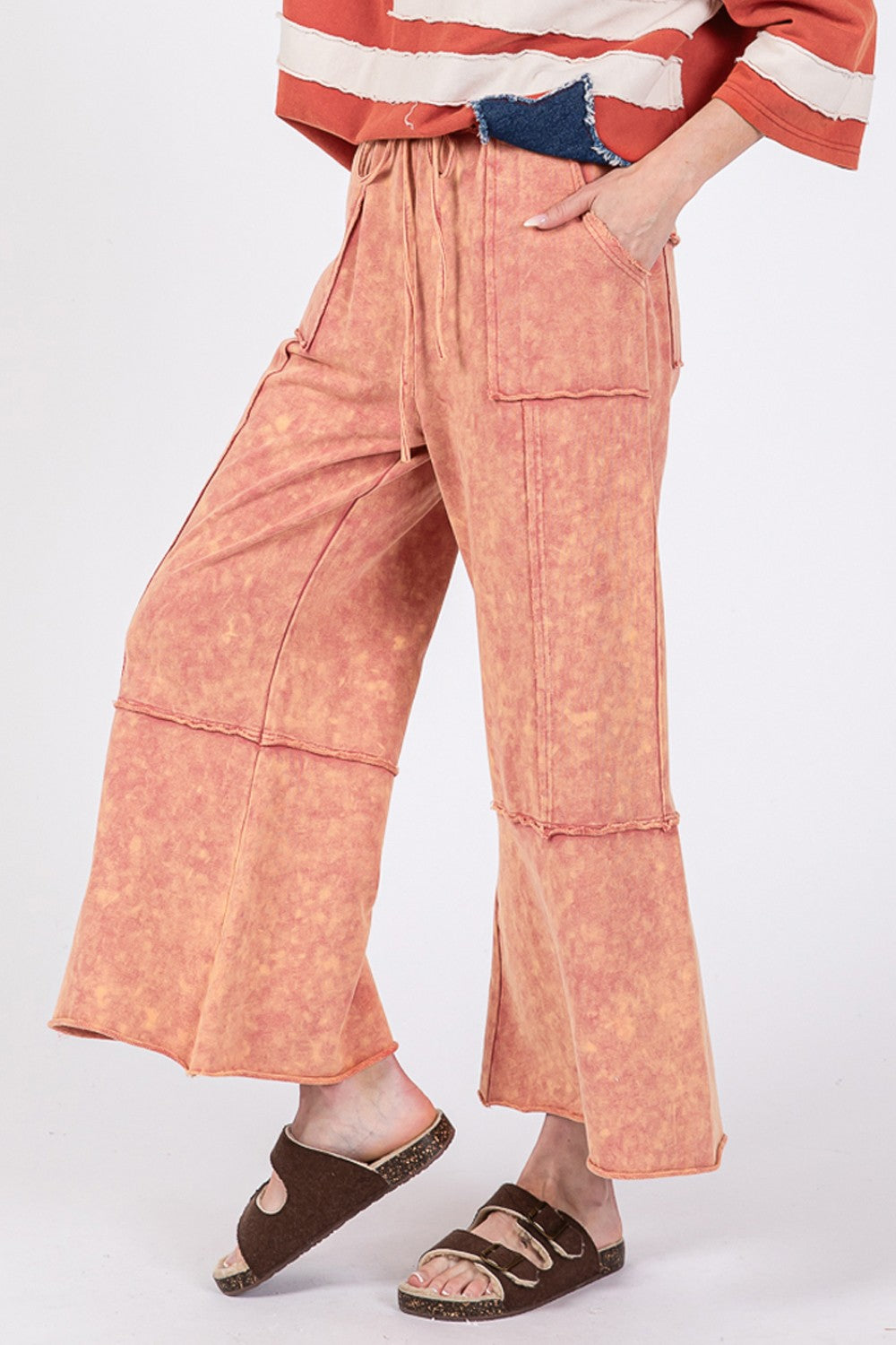 Person wearing SAGE + FIG Mineral Washed Terry Wide Leg Pants with an adjustable waistband and oversized pockets in orange, paired with sandals and a striped top.