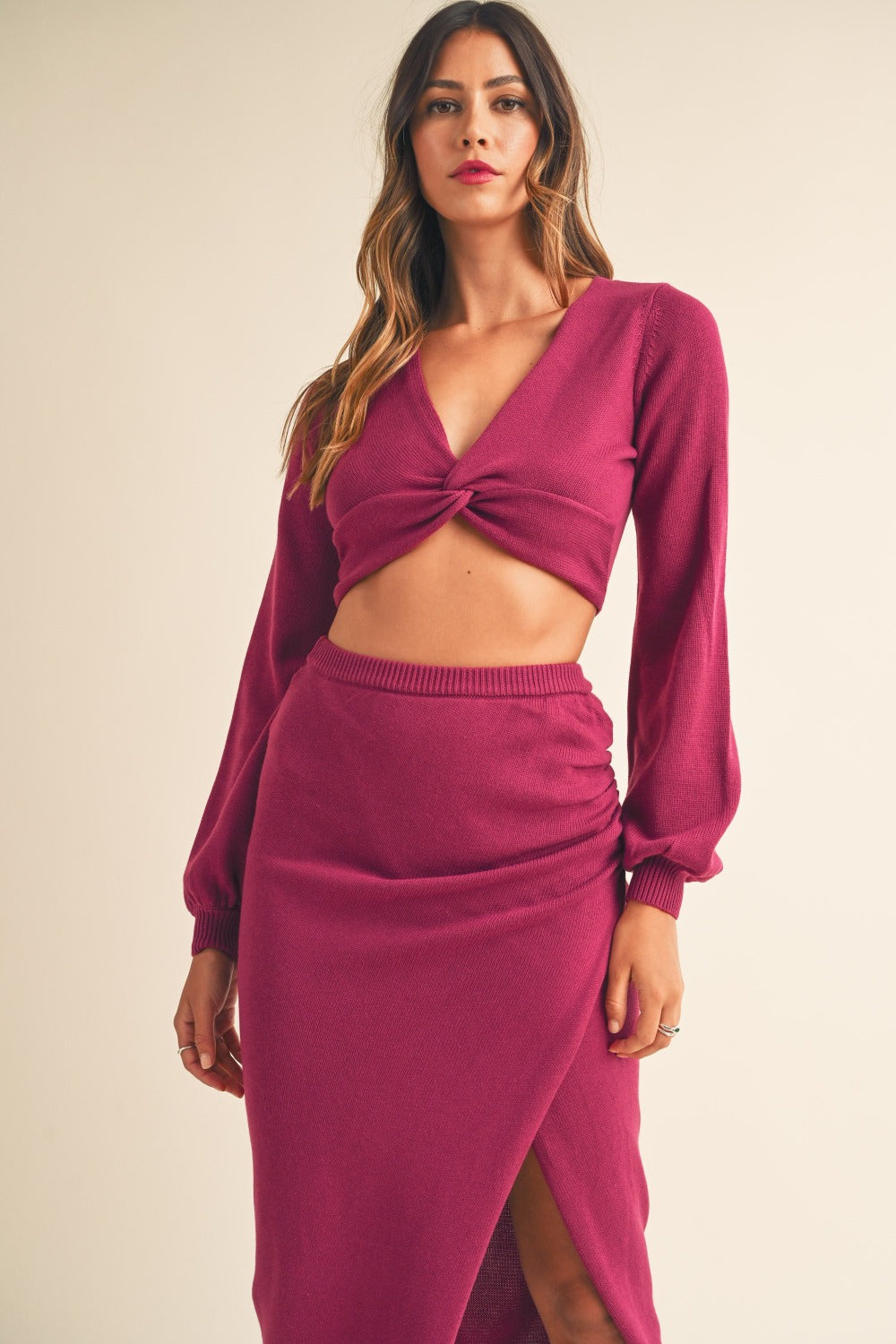 A woman wearing the MABLE Front Twisted Knit Top and Midi Skirt Set, featuring a long-sleeved, magenta knitted top with a front twist detail and a matching high-waisted, thigh-slit midi skirt, stands against a plain background.