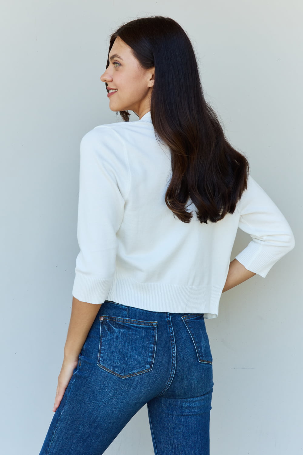 A stylish person wearing sunglasses, a Doublju My Favorite Full Size 3/4 Sleeve Cropped Cardigan in Ivory, and blue jeans poses in front of a plain background, effortlessly highlighting their versatile look.