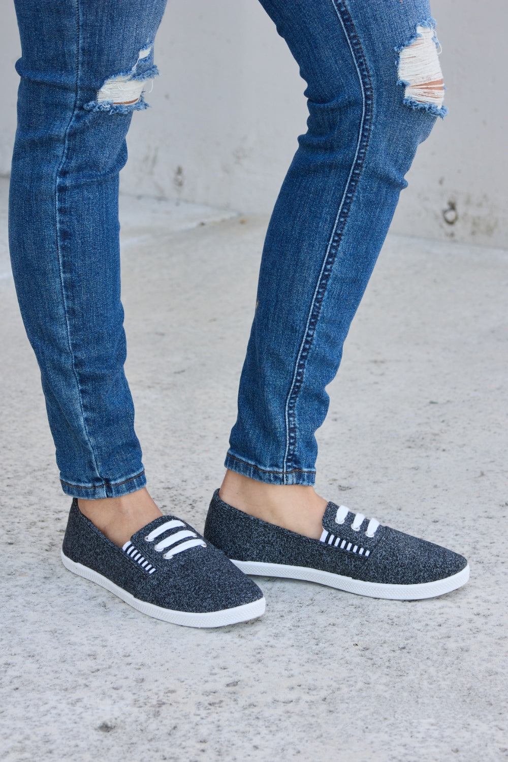 A person wearing ripped blue jeans and the fashionable Forever Link Round Toe Slip-On Flat Sneakers with white elastic bands.