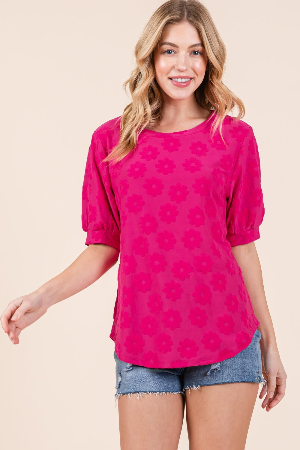 A woman showcases her versatile style in the BOMBOM Textured Floral Pattern Top, with its bright pink hue and feminine design, paired effortlessly with denim shorts against a plain background.