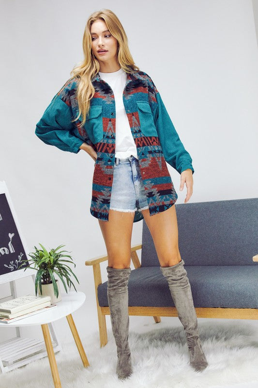 A woman with long blonde hair wears a blue and gray patterned shirt over a white crop top and dark jeans, standing indoors near a window, her casual **Printed Button Down Long Sleeve Jacket** draped casually over her shoulders.