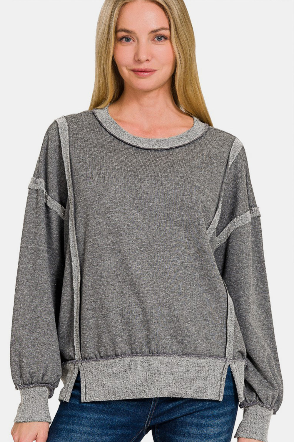 A woman with long blonde hair is seen in a neutral pose against a plain white background, wearing the grey Zenana Washed Exposed-Seam Sweatshirt featuring a crew neck and ribbed hem. Her casual wear is perfect for layering in any season.