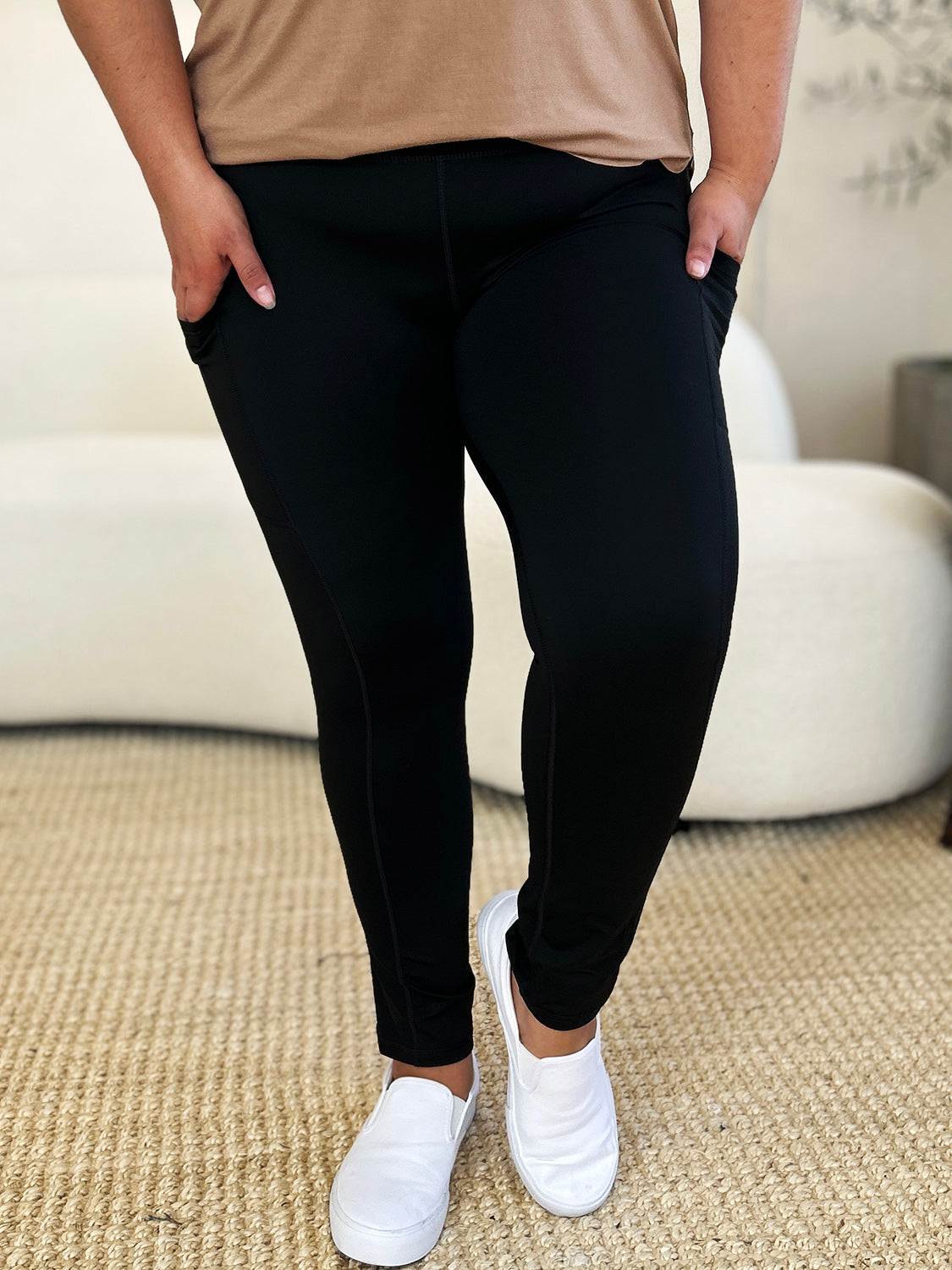 Person wearing Wide Waistband Sports Leggings made of stretchy nylon spandex and white slip-on shoes, standing on a beige carpet with a white couch in the background.