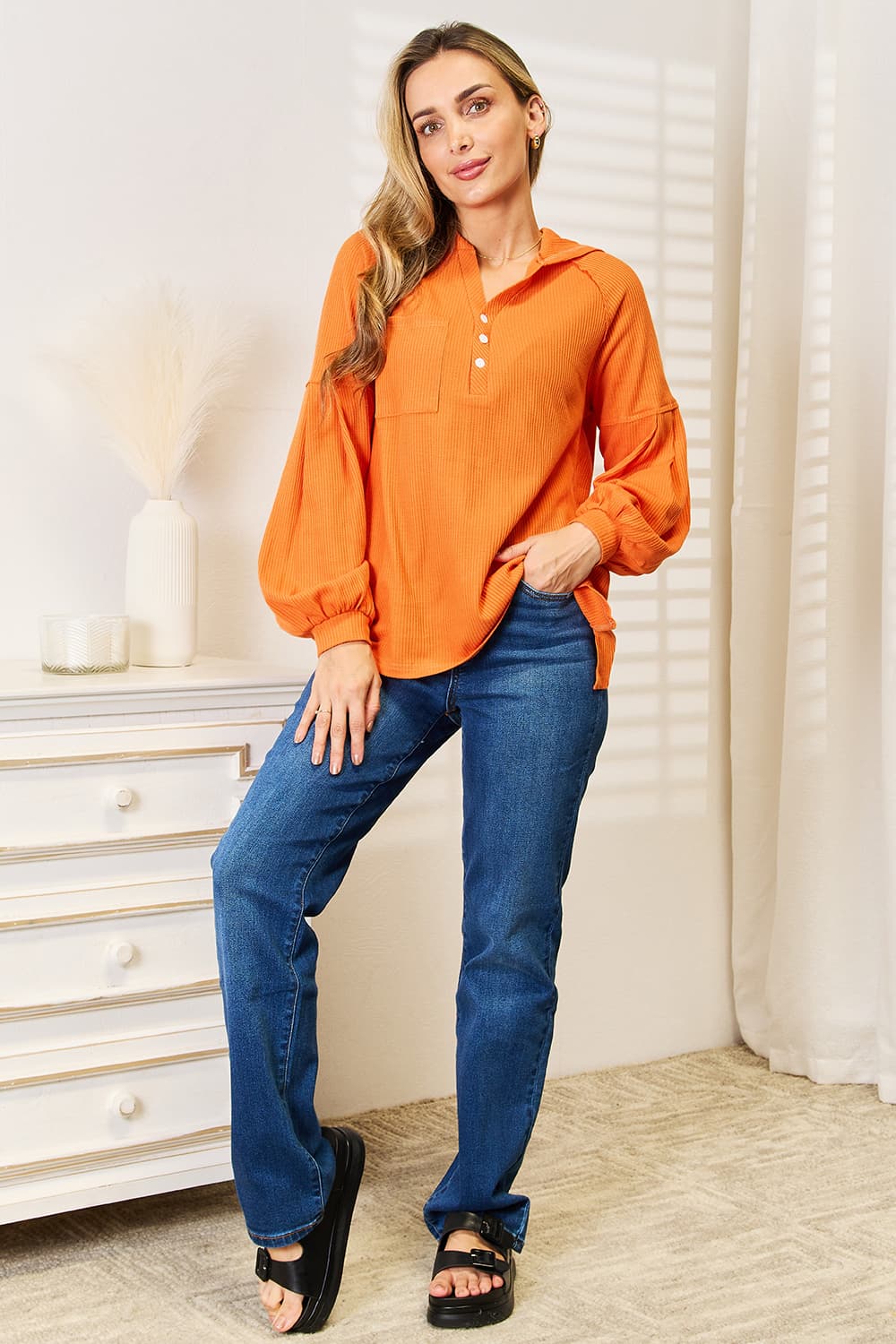 A person wearing an orange Basic Bae Side Slit Buttoned Waffle Knit Hoodie with blue jeans stands indoors near a white dresser, smiling and looking to the side.