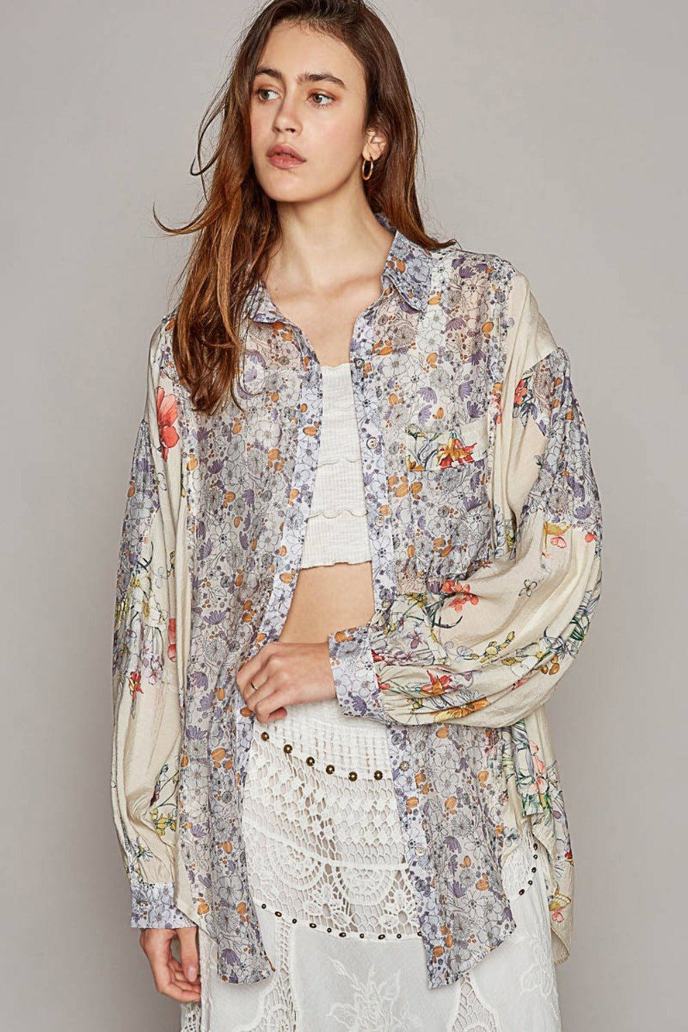 Wearing the POL Long Sleeve Floral Button Down Shirt and a layered skirt, a person stands outdoors under a wooden structure surrounded by plants, with mountains in the background.