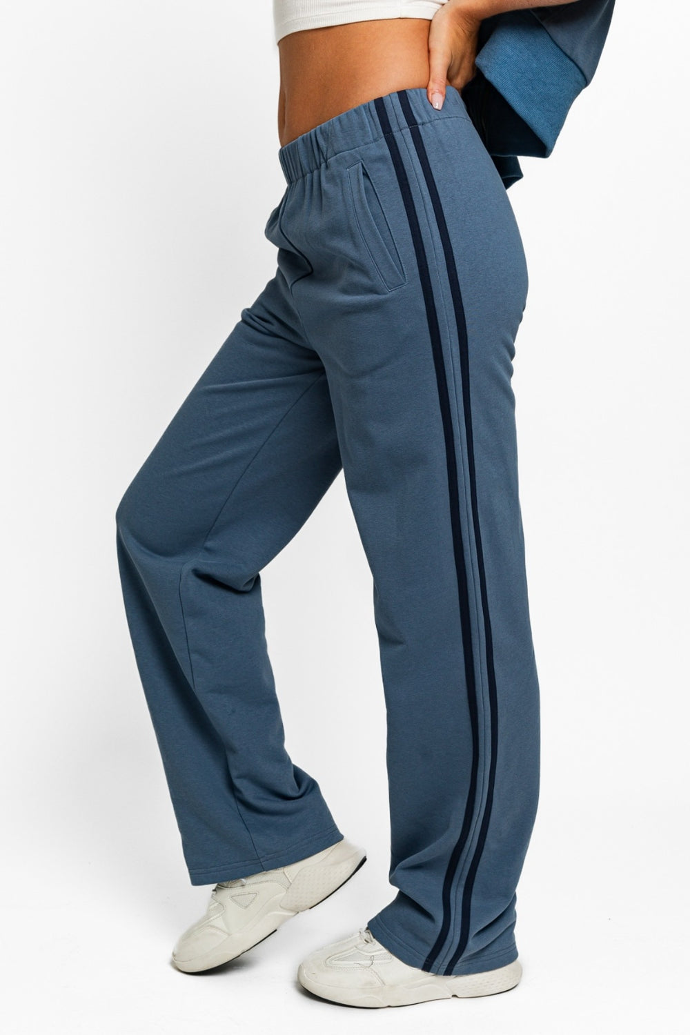 A person is shown wearing the Le Lis High Waisted Side Stripes Straight Track Sweatpants, hands in pockets, with a partial view from the torso down. The blue jogger pants feature striking black side stripes and are styled with a beige crop top and white shoes, perfectly capturing the athleisure aesthetic.