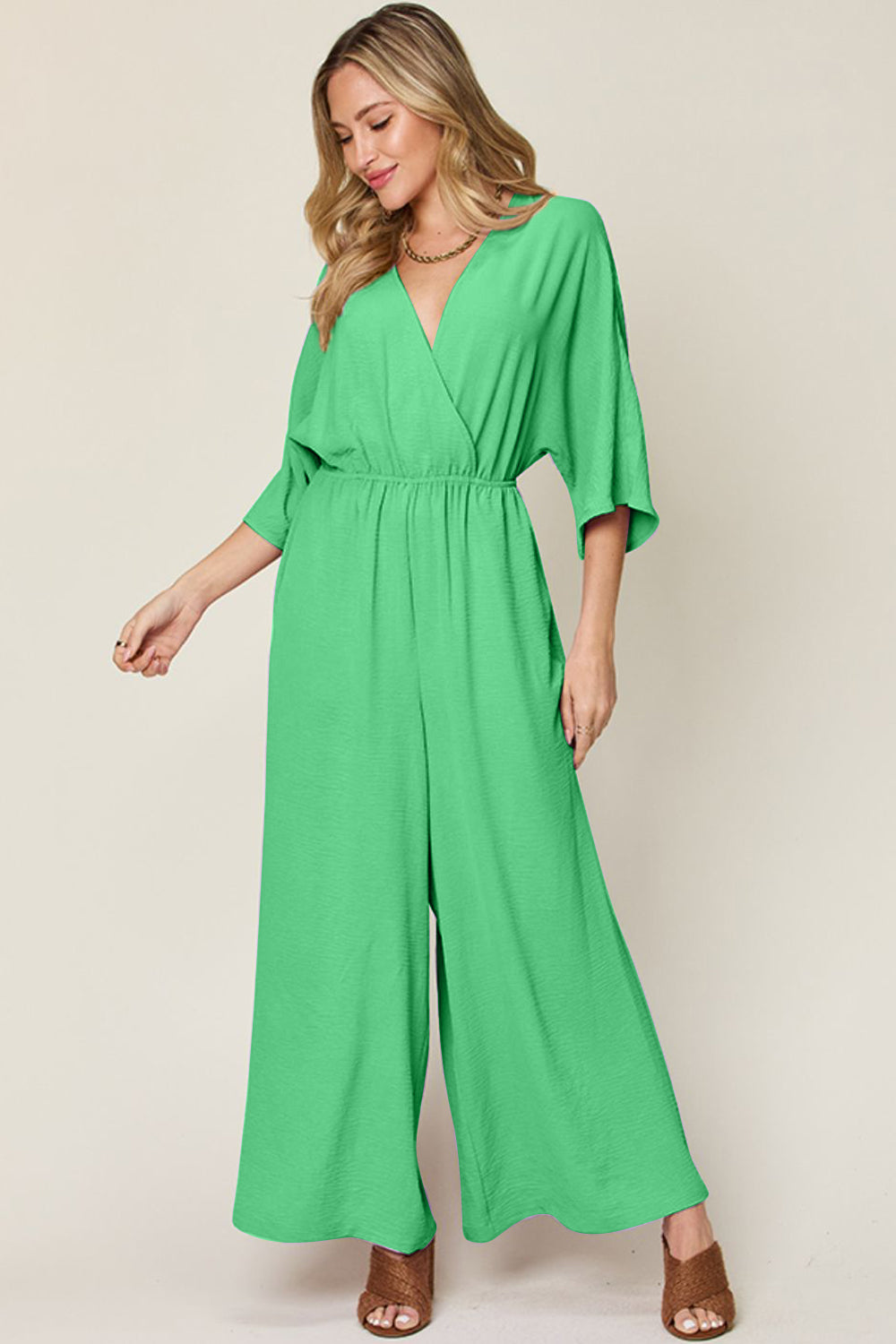 Person wearing the Double Take Full Size Surplice Wide Leg Jumpsuit with Pockets in royal blue, featuring wide legs, a deep V-neckline, and ruched detailing, posing with hands on hips against a plain light-colored background.