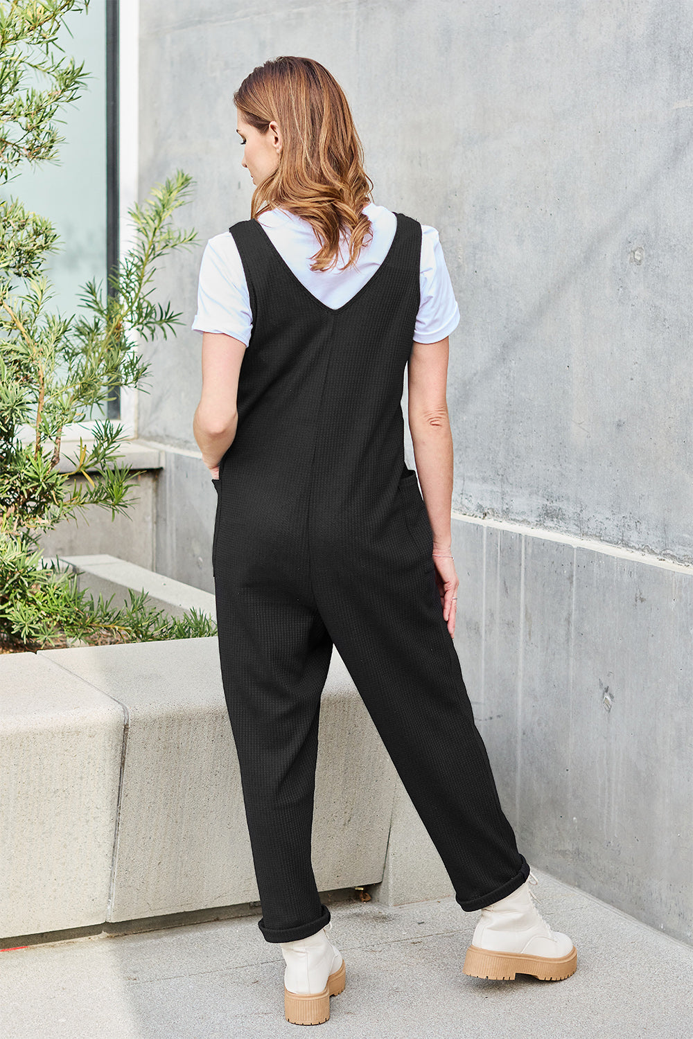 A person stands outdoors wearing the Double Take Full Size Sleeveless Straight Jumpsuit in blue and brown sandals, holding a cream-colored bag. In the background, there's a wooden bench and palm trees.