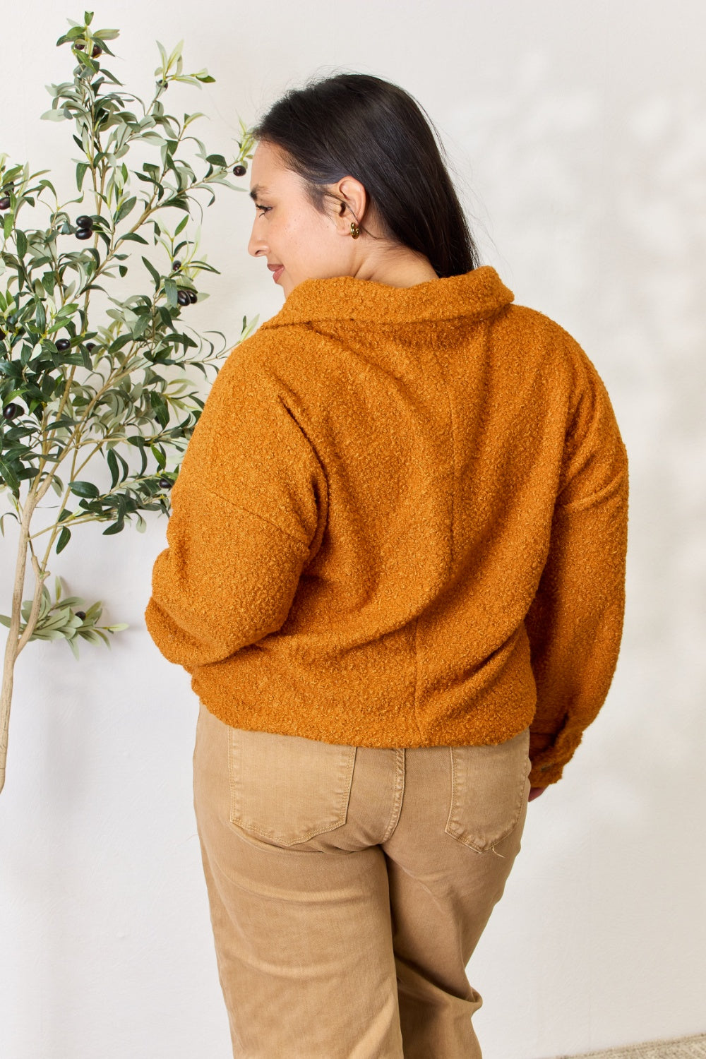 A person wears a brown textured Culture Code Full Size Half Button Turtleneck Sweatshirt made from breathable fabrics and tan pants, standing next to a leafy plant against a light background.