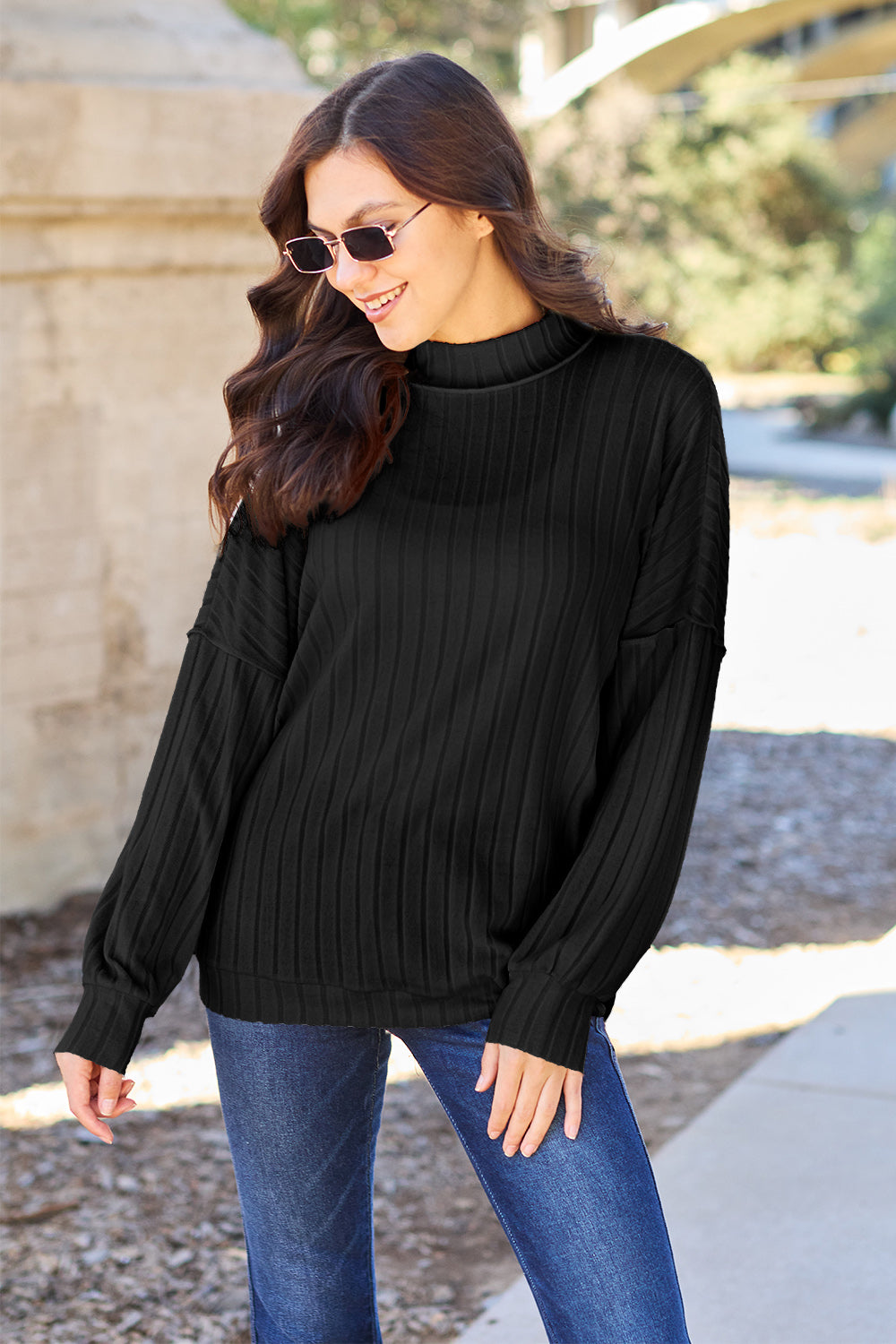 A woman with long hair is wearing a green Basic Bae Full Size Ribbed Exposed Seam Mock Neck Knit Top, blue jeans, and sunglasses. She is standing outdoors and smiling.