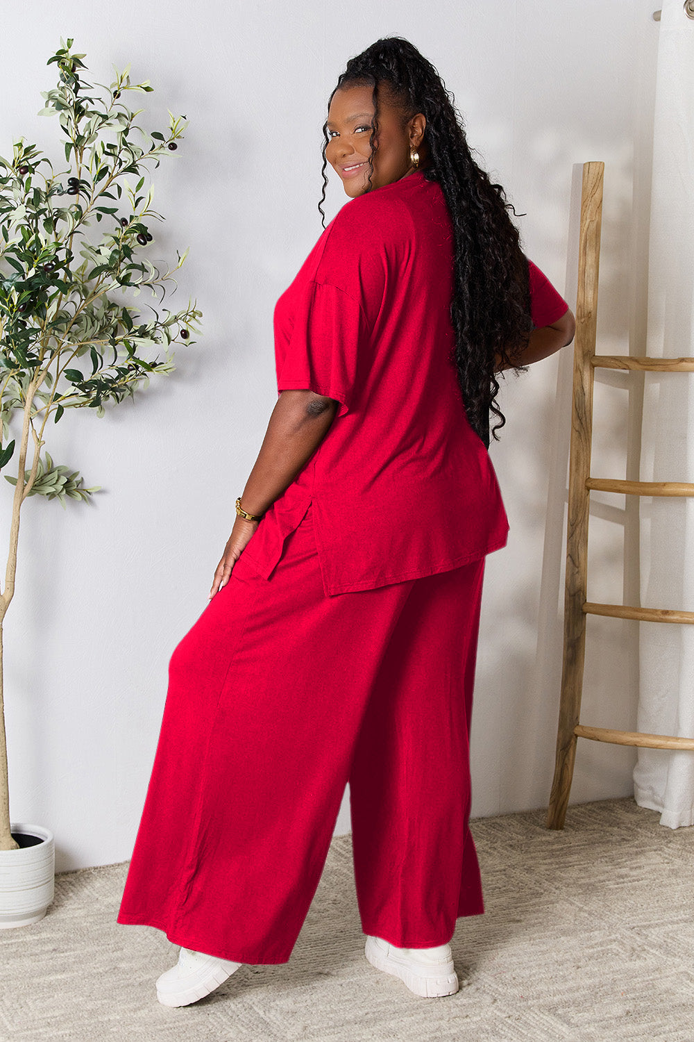 A woman stands near a potted plant and wooden ladder, wearing the Double Take Full Size Round Neck Slit Top and Pants Set in a bright pink two-piece outfit with matching wide-leg pants that are available in plus size and are easily machine washable.