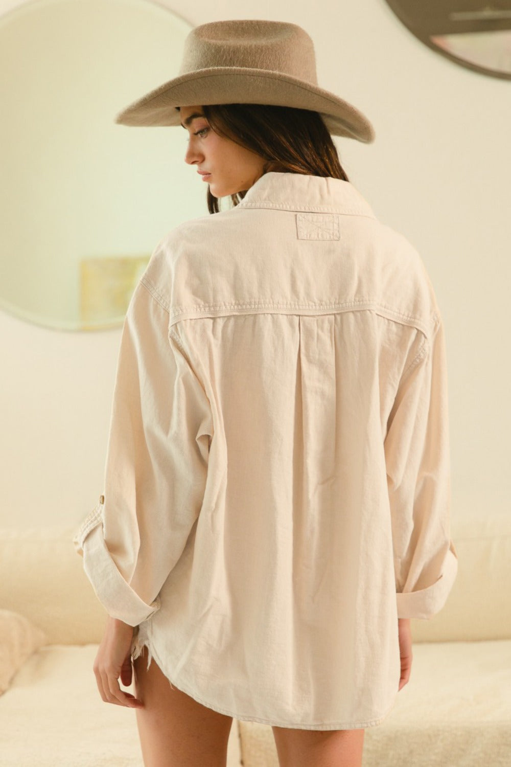 A person wearing the BiBi Button Down Stitch Detail Shirt with large chest pockets, denim shorts, and a beige hat stands indoors smiling.