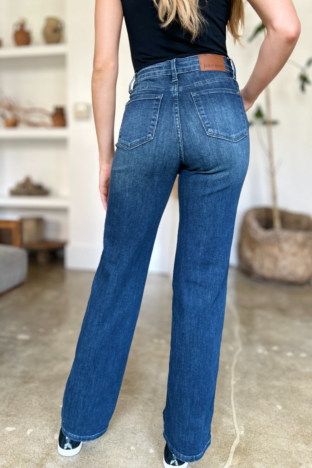 A person wearing a black top and Judy Blue Full Size Tummy Control Straight Jeans stands indoors with one hand on their hip. Their shoes are black with white soles. A plant and shelves are in the background.