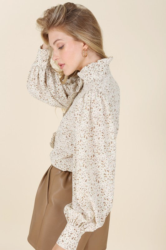 A woman wearing a stand collar floral frill blouse with puff sleeves and tan pants is standing against a beige background, looking to the side.