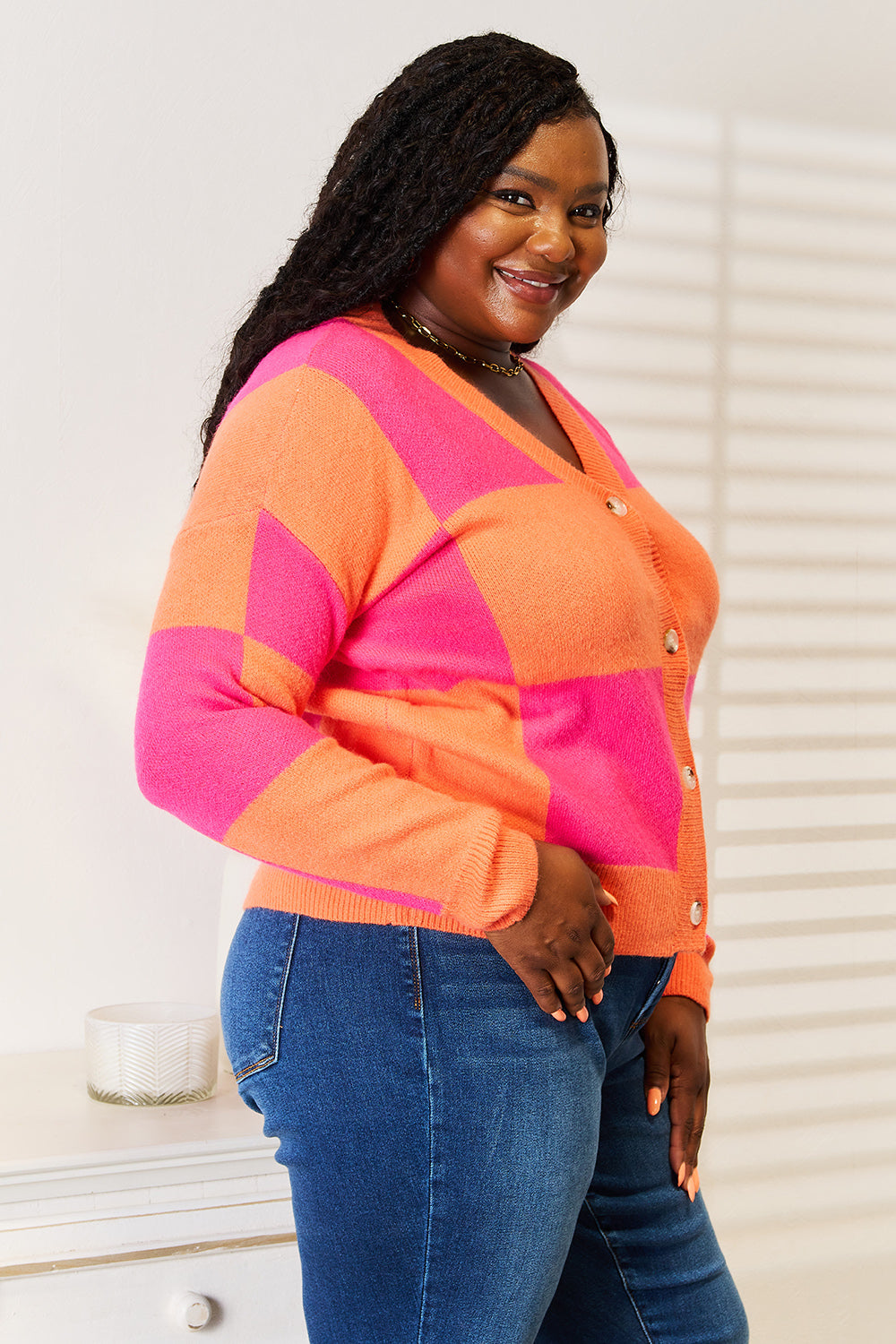 A woman with long hair stands smiling, wearing the Angel Wings Woven Right Checkered V-Neck Dropped Shoulder Cardigan in pink and orange over blue jeans.