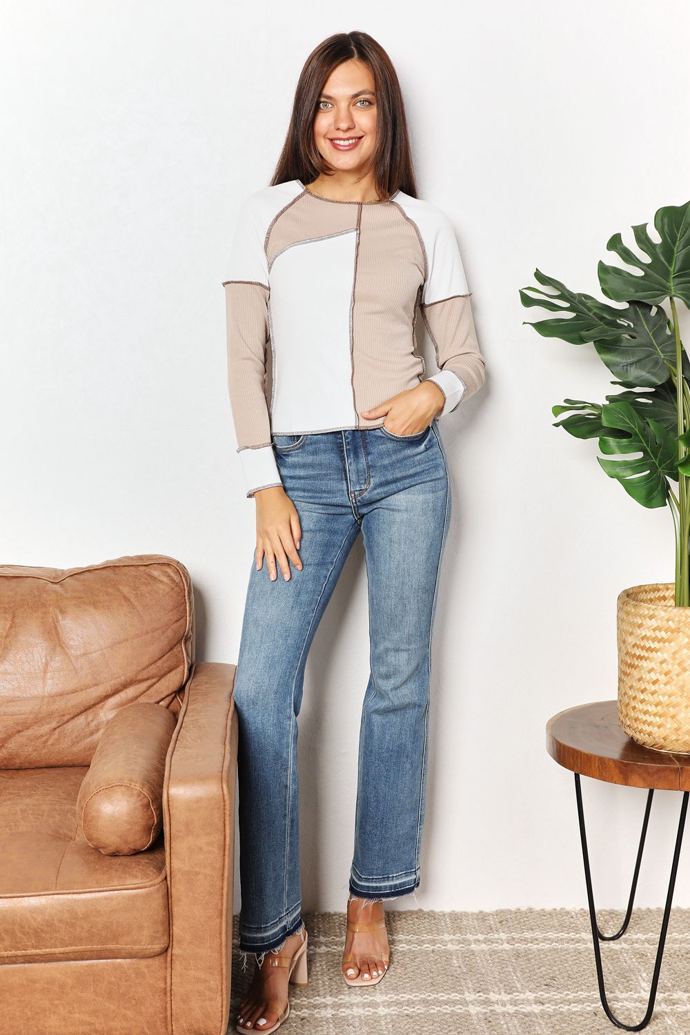 A woman with long brown hair is wearing the "Double Take Color Block Exposed Seam Top" in beige and white, paired with jeans. She stands with one hand in her pocket and the other touching her hair, smiling. A plant is visible in the background.