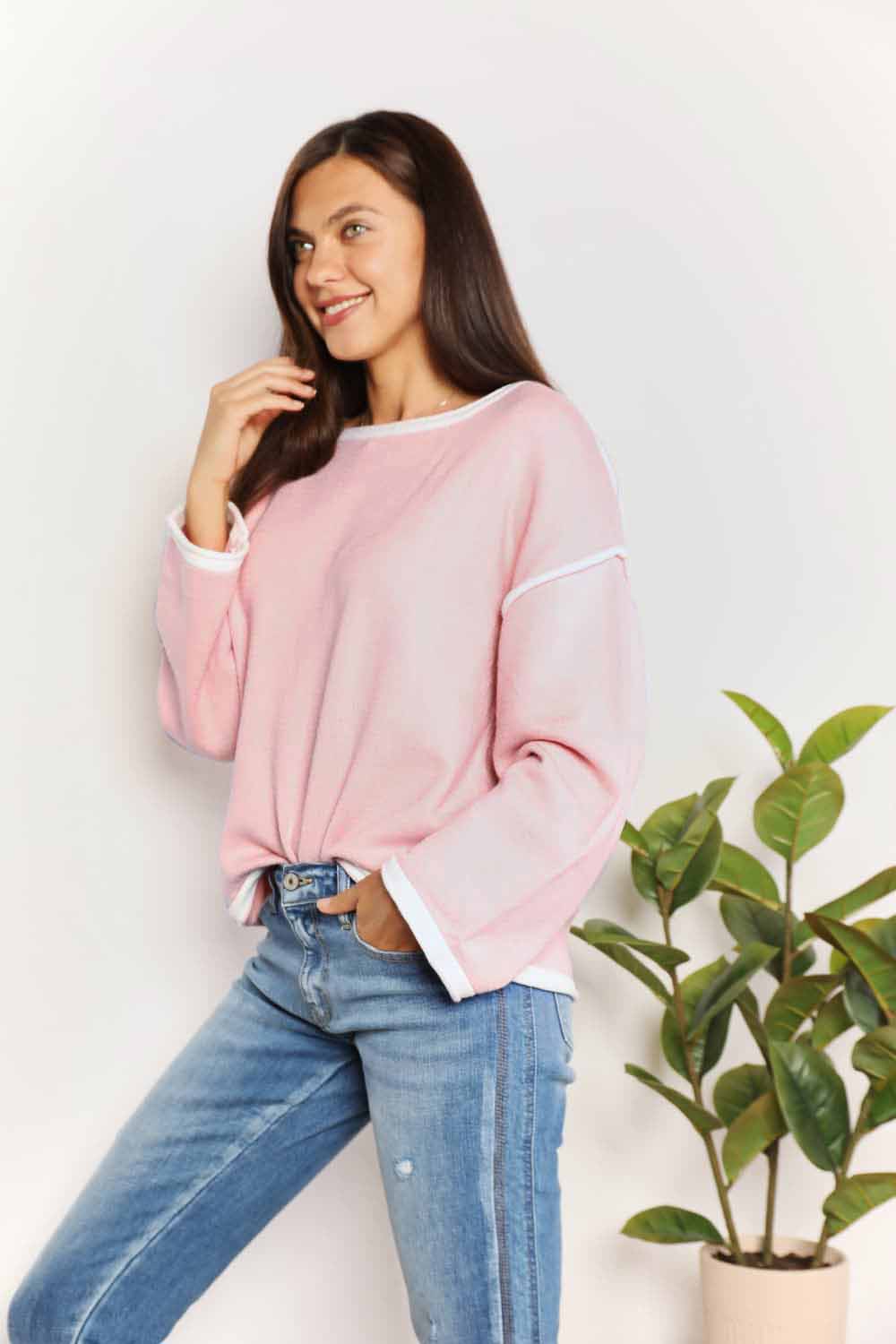 A woman with long curly hair is wearing the Angel Wings Contrast Detail Dropped Shoulder Knit Top in pink, paired with blue jeans, standing against a light background.