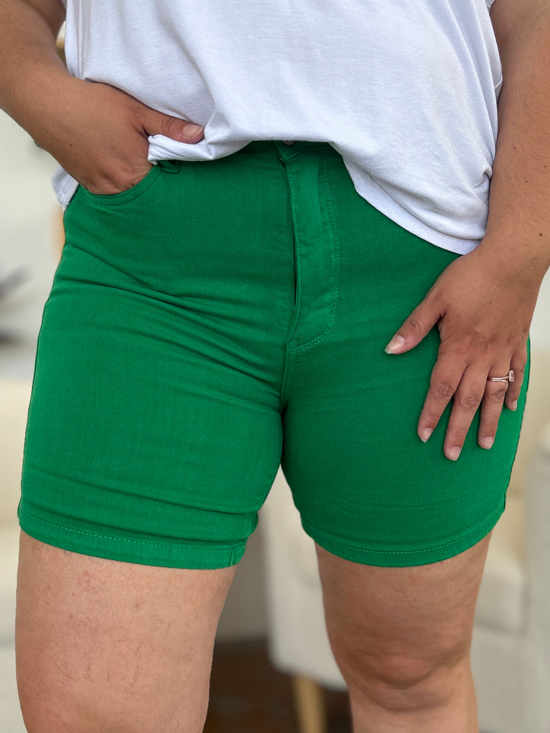 Two women stand indoors wearing white tops and Judy Blue Full Size Tummy Control Garment Dyed Denim Shorts in green. The woman on the left has her hand in her pocket, and both are smiling. There are plants and decorations in the background.