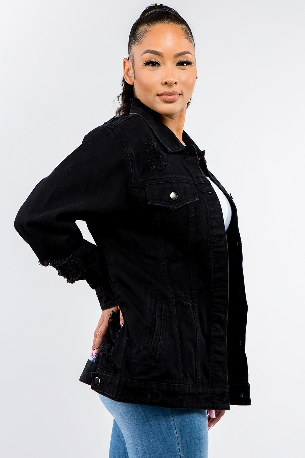 A woman stands against a plain background, epitomizing casual outfits with a touch of vintage look, wearing the American Bazi Full Size Button Up Distressed Denim Jacket over a white crop top and jeans.