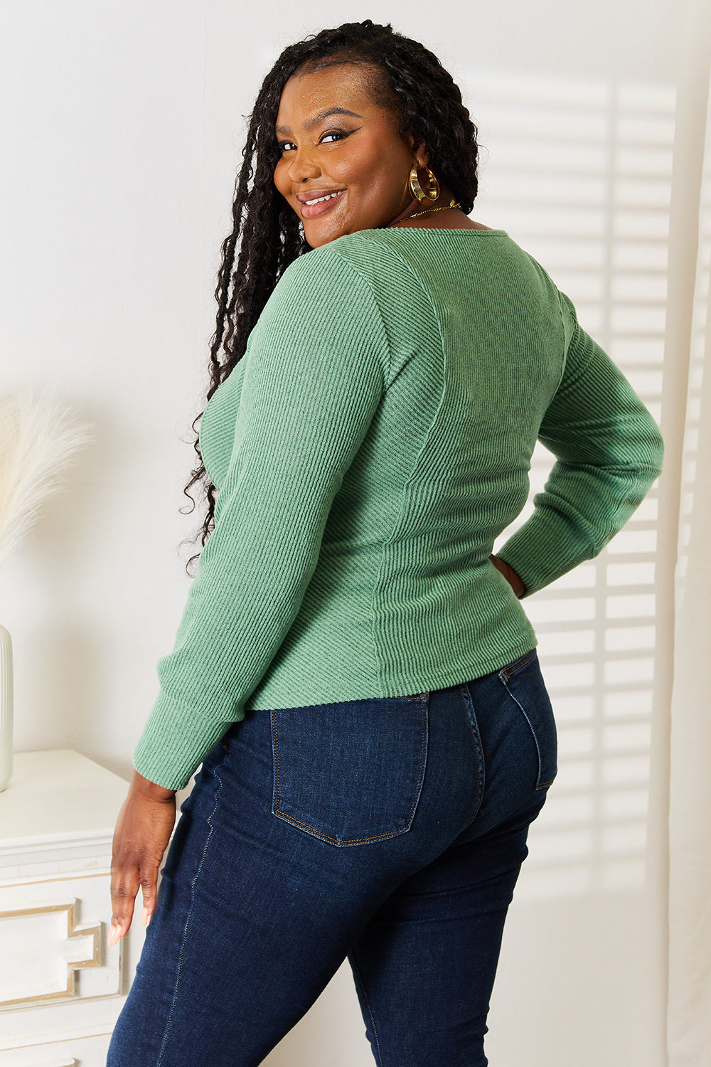 A person wearing a fashion-forward Basic Bae Scoop Neck Long Sleeve Top in green and dark jeans stands indoors, smiling and looking to the side.