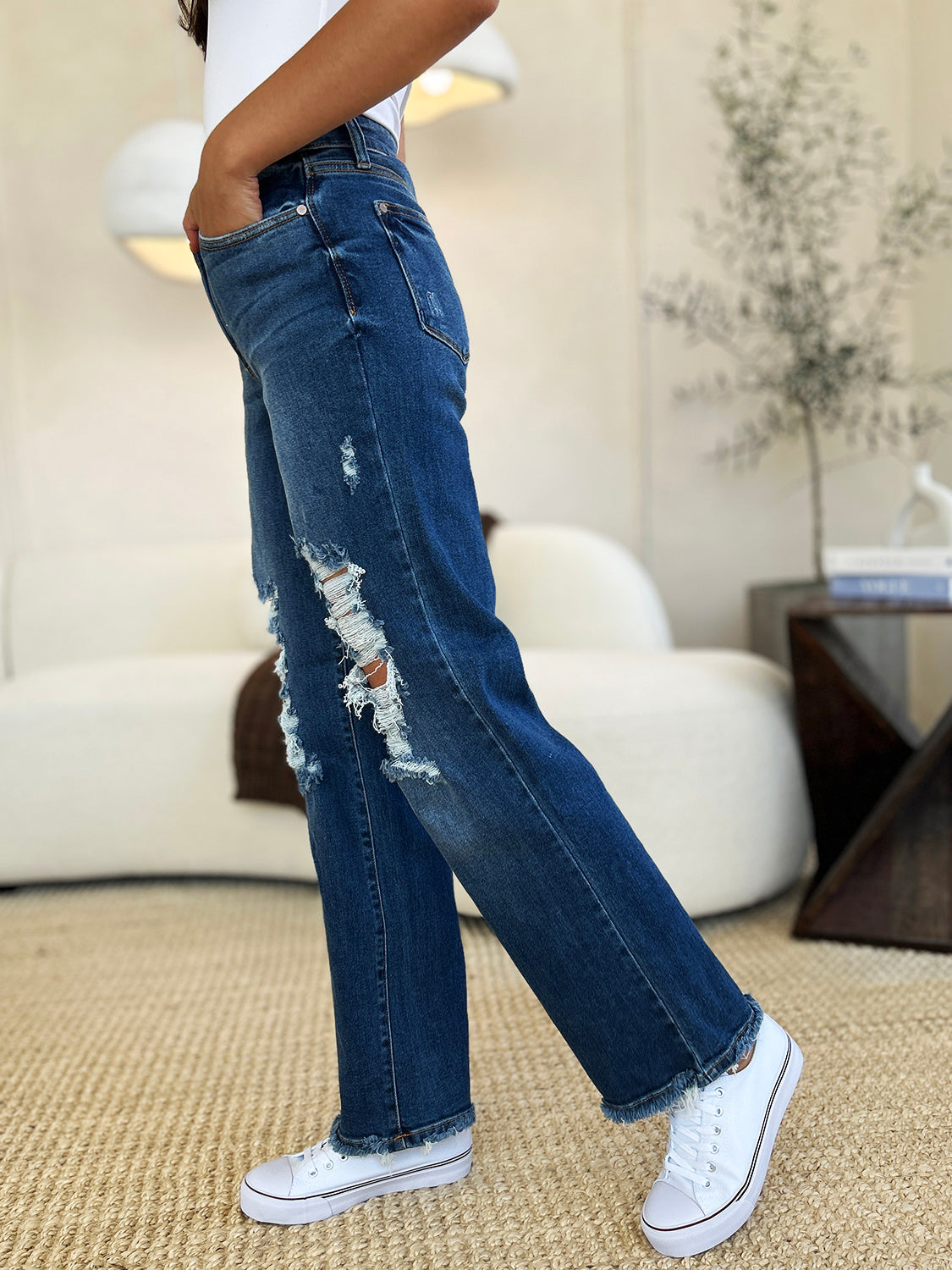 Person standing indoors wearing Judy Blue Full Size Mid Rise Distressed Raw Hem Jeans and black sneakers.